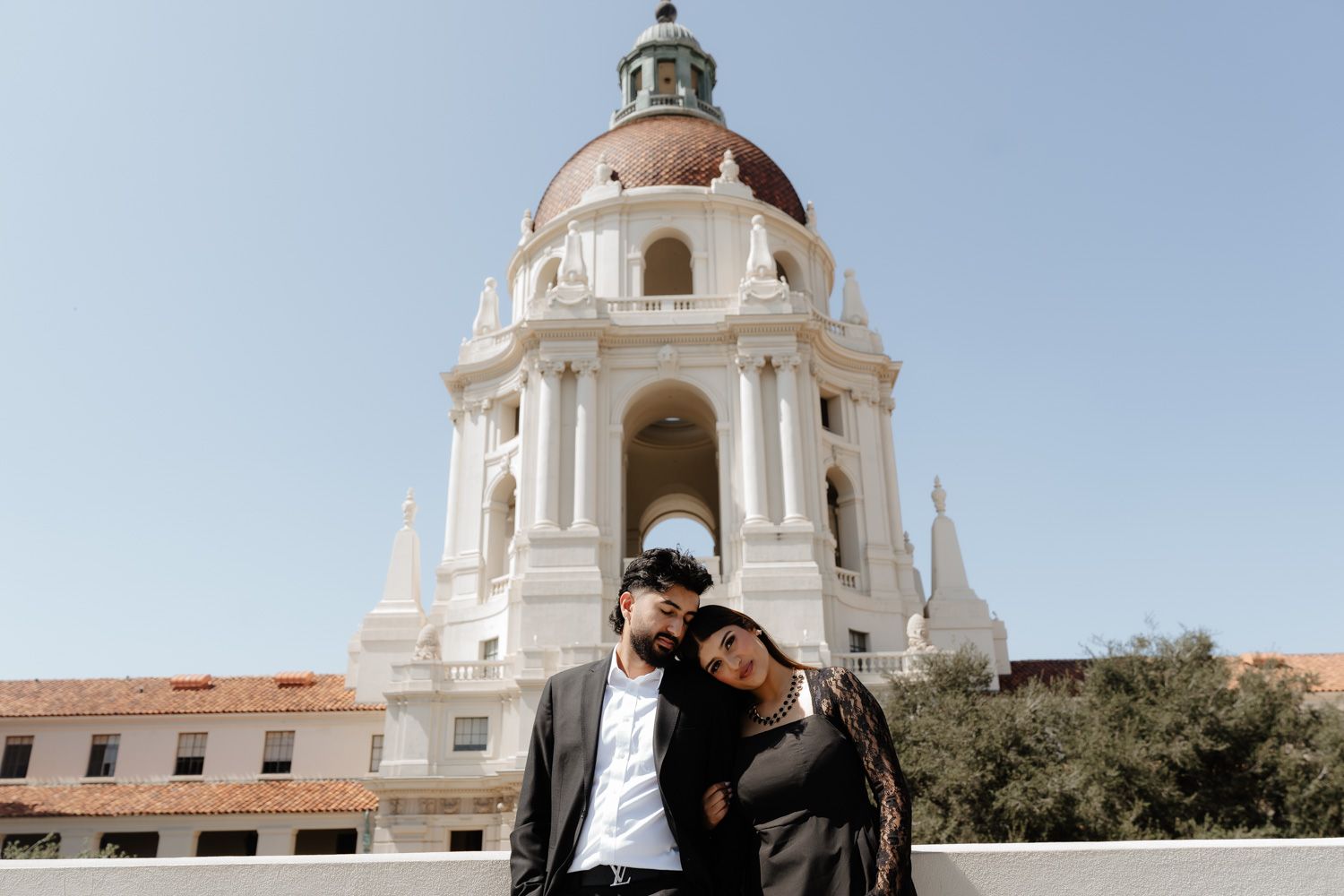 Couple posing in front of Pasadena City Hall dome, woman leaning on mans shoulder, both in black outfits.