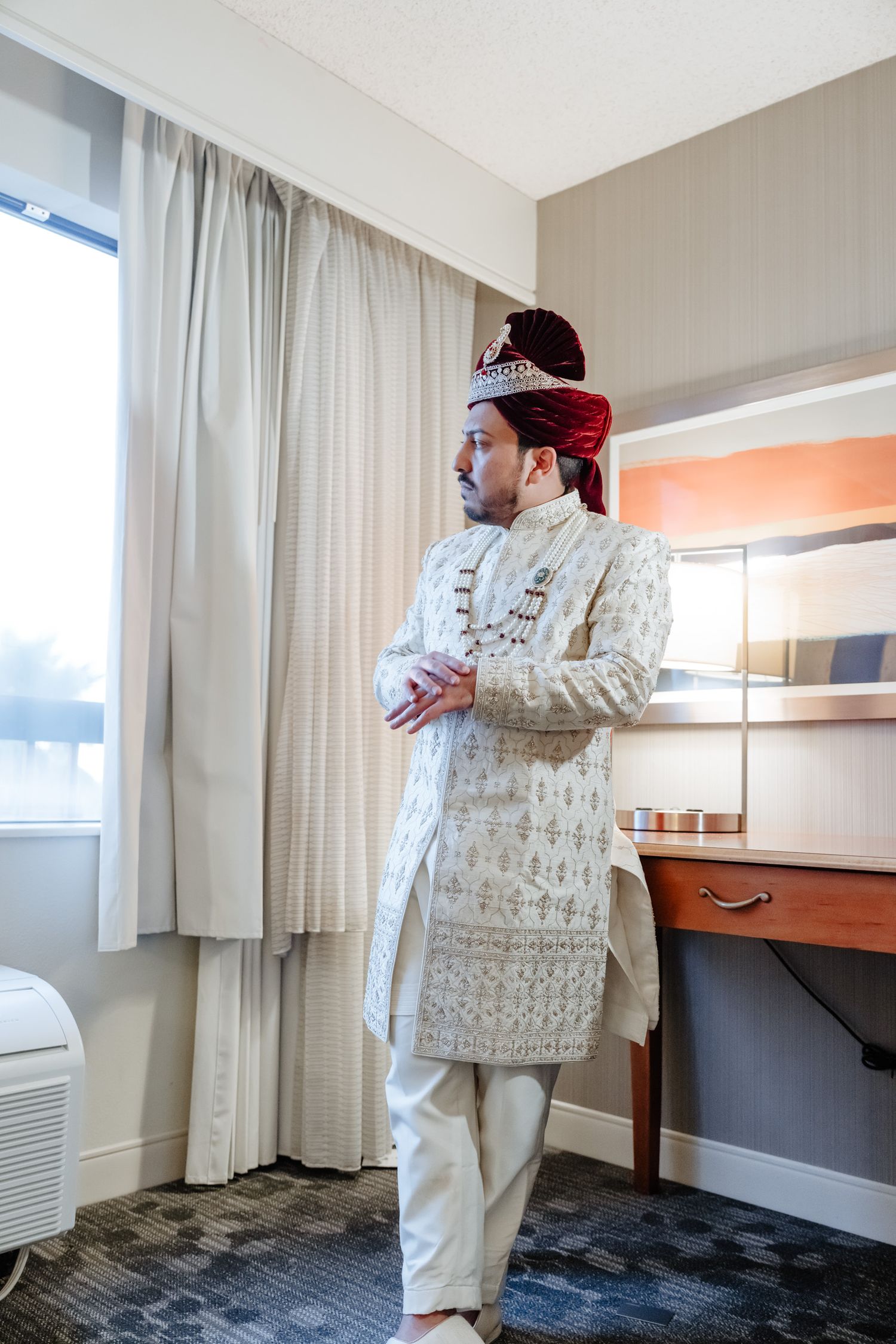 A person in traditional South Asian wedding attire with a white embroidered sherwani and red turban stands in a hotel room.