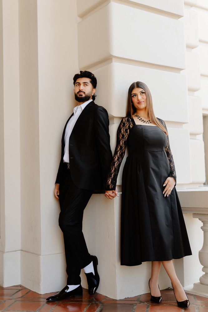 Elegant couple in black formal attire holding hands while leaning against white architectural column.
