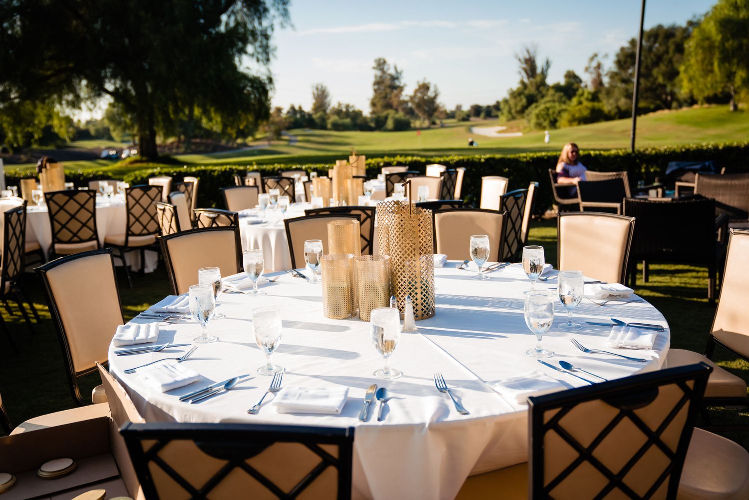 Elegant outdoor wedding reception tables set with white linens and floral centerpieces under trees at sunset.