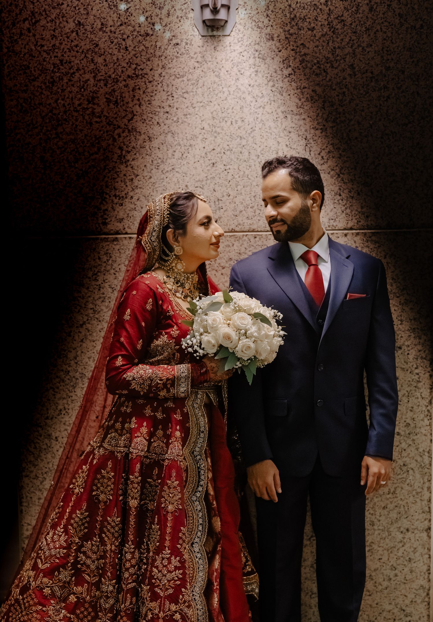 A couple in traditional South Asian wedding attire, with the bride in a red lehenga and groom in dark suit, share romantic moments.