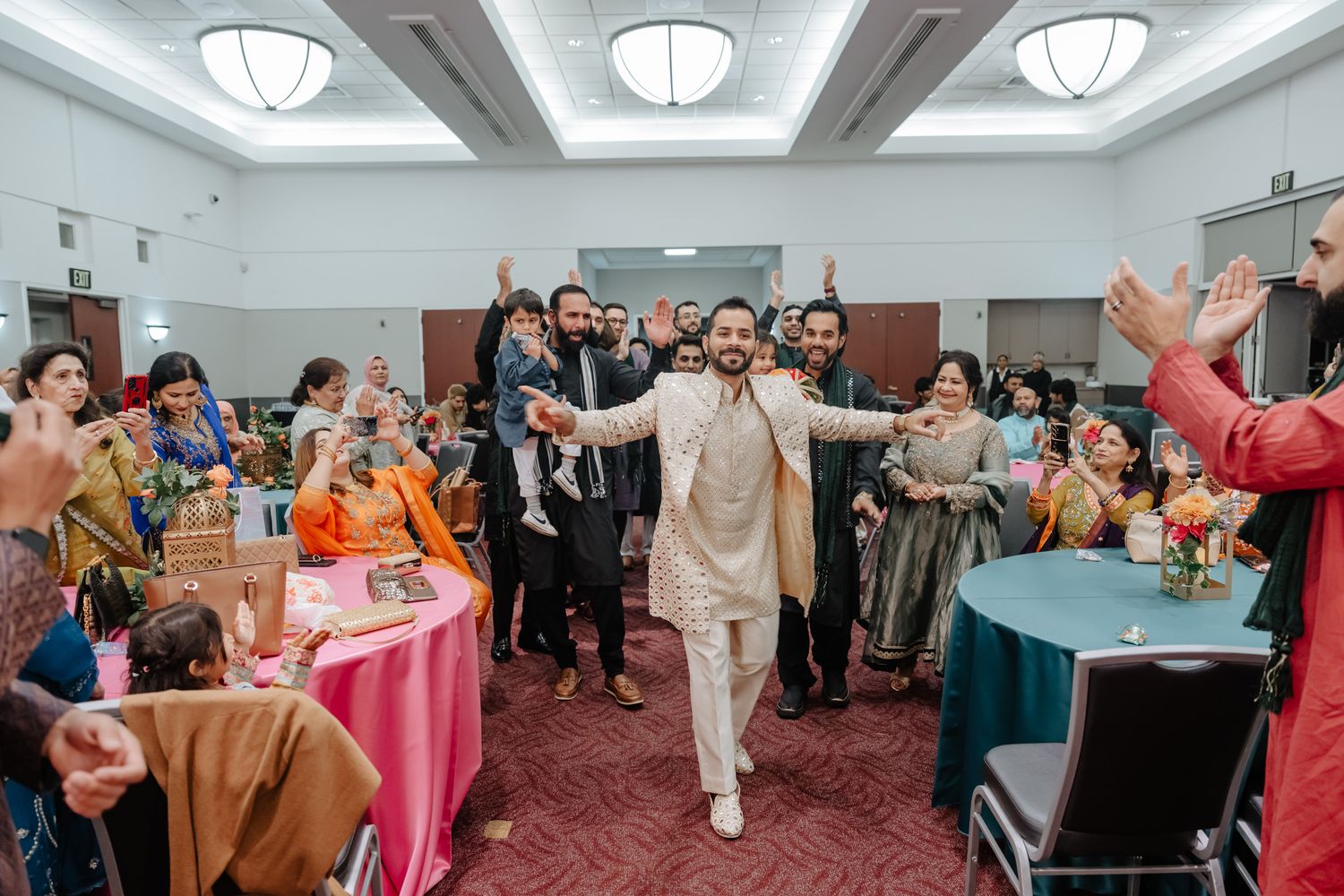 Wedding guests dance and celebrate on a carpeted reception hall floor under decorative dome lighting fixtures.