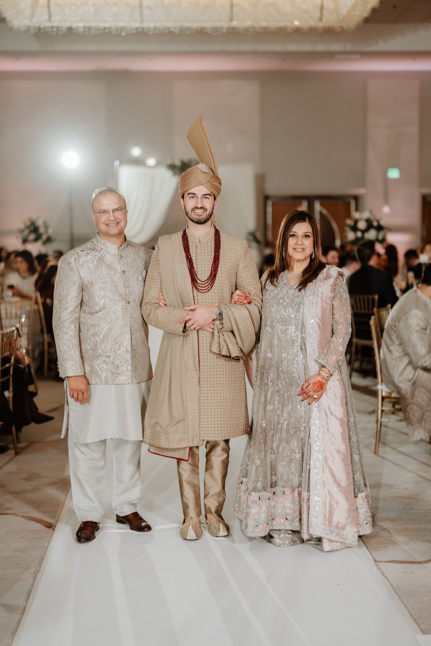 Wedding party group poses together in formal attire against a light-colored backdrop.