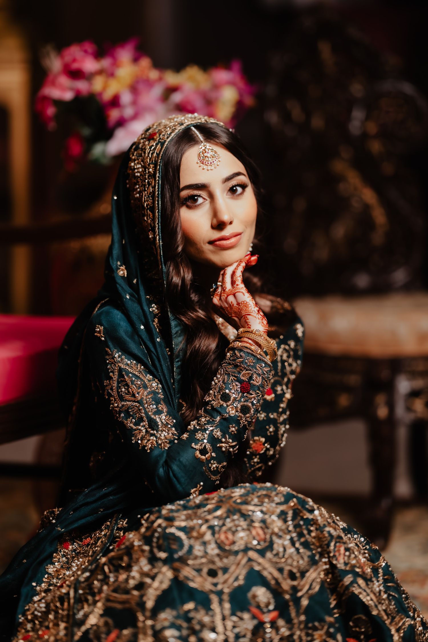 Elegant traditional bridal portrait showcasing ornate embroidered dress, jewelry and henna details in dark lighting.