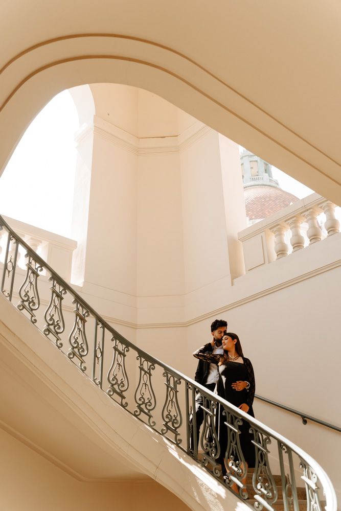 Elegant couple standing on ornate spiral staircase in a grand white classical building interior.
