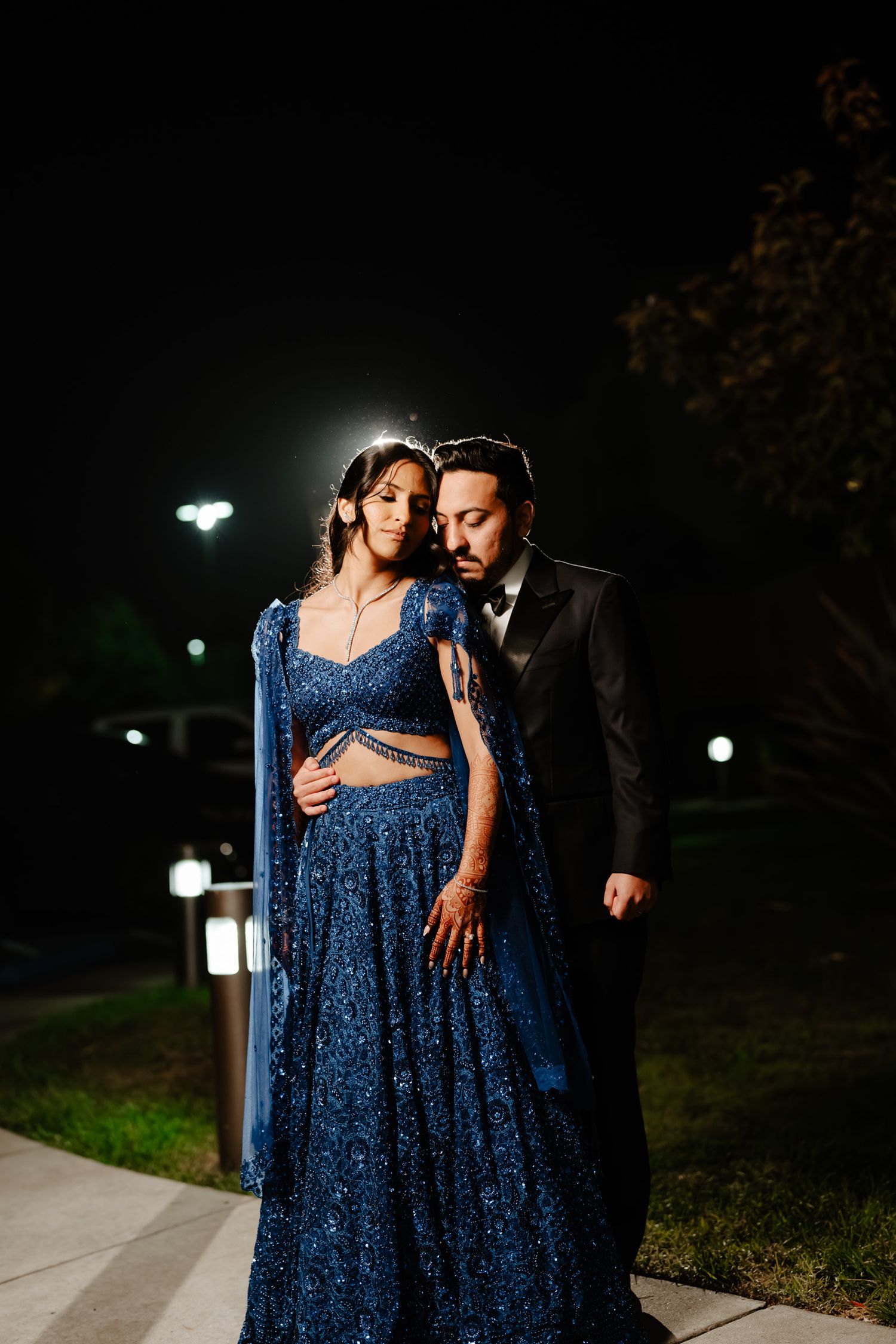 Couple in formal attire pose romantically at night with blue sequined dress and dramatic lighting.