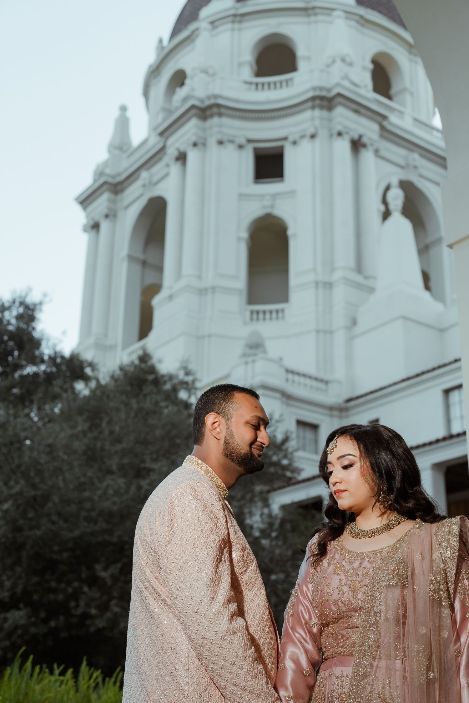 A couple wearing matching pink traditional Indian outfits stand in front of a white church tower.
