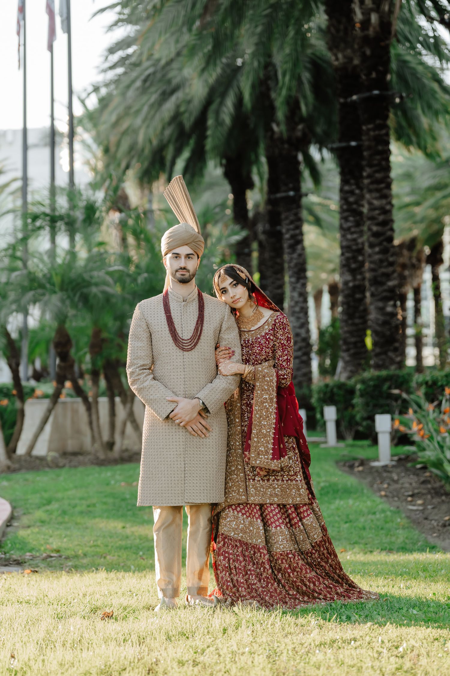 A couple in traditional wedding attire poses outdoors among palm trees on a grassy lawn in natural lighting.
