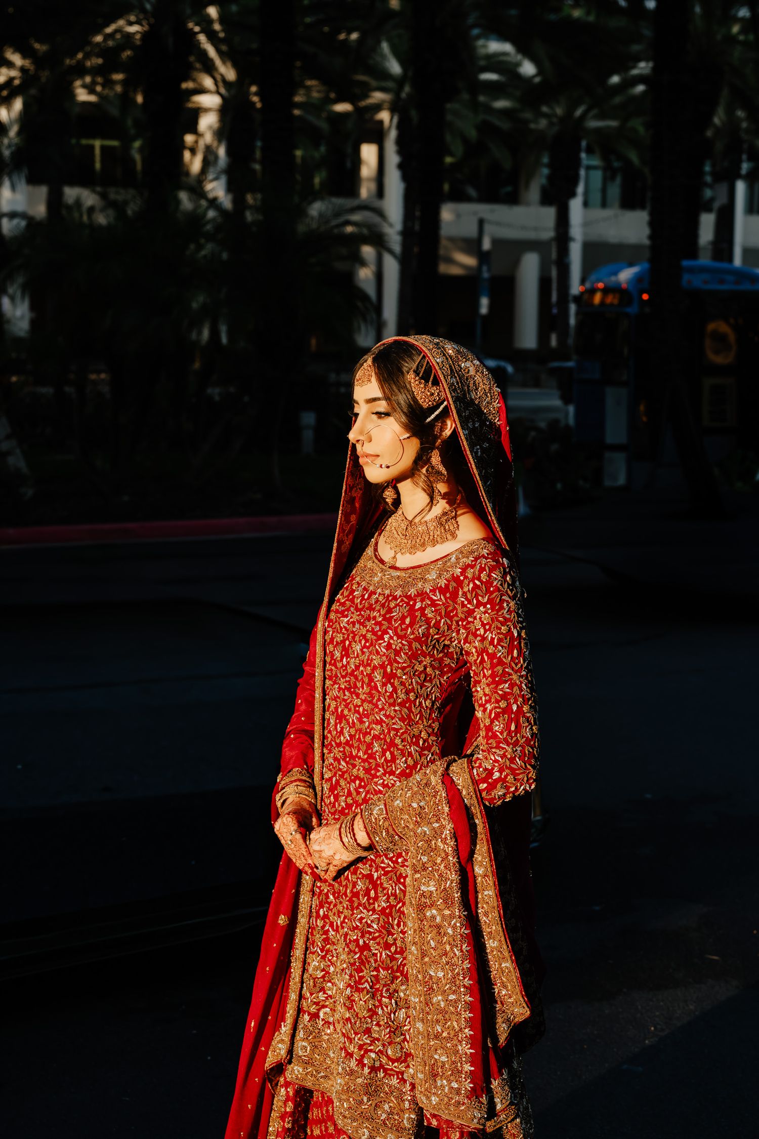 A bride in a stunning red and gold embroidered lehenga poses outdoors at sunset.