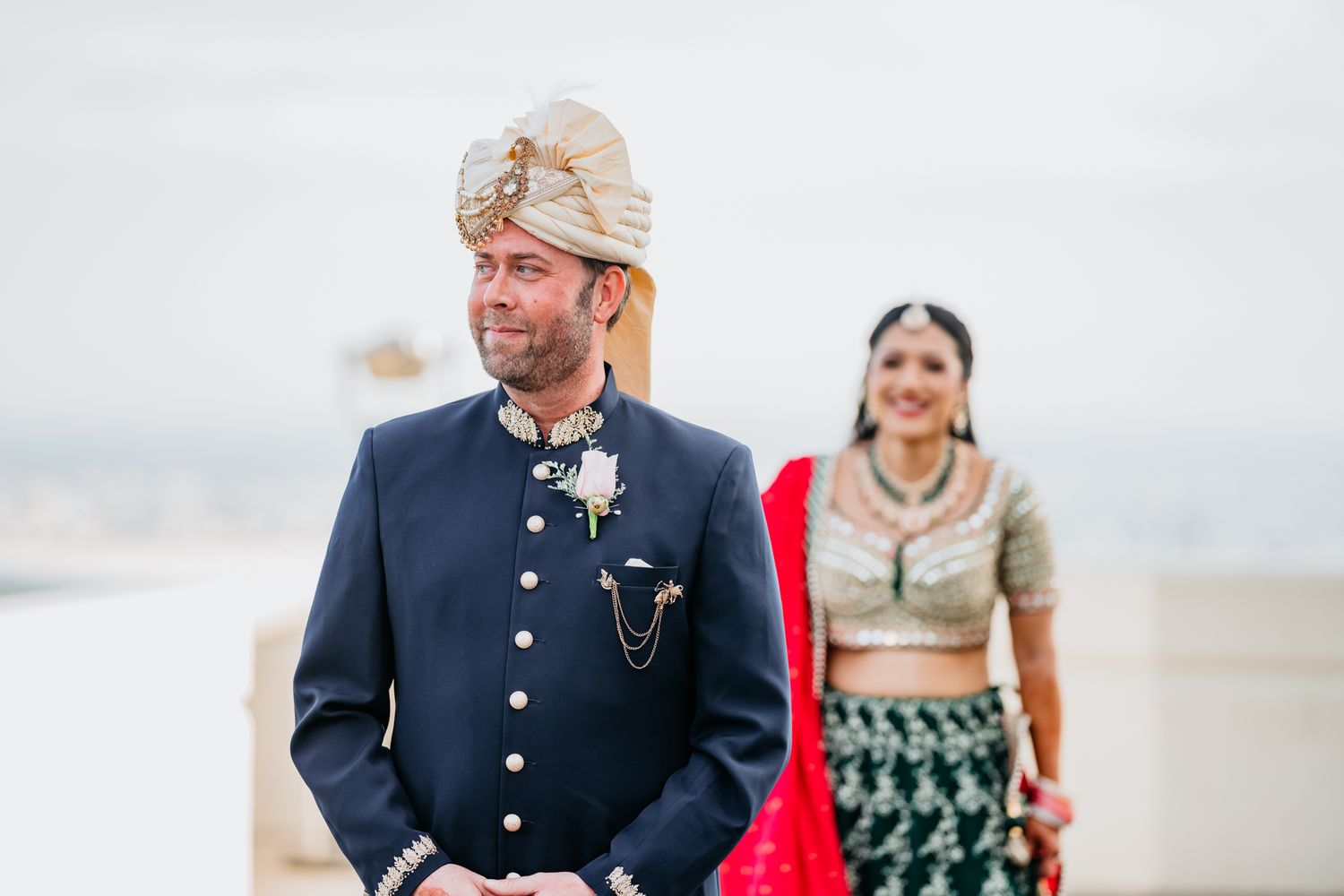 A couple in traditional Indian wedding attire stand together on a beach with cloudy skies in the background.