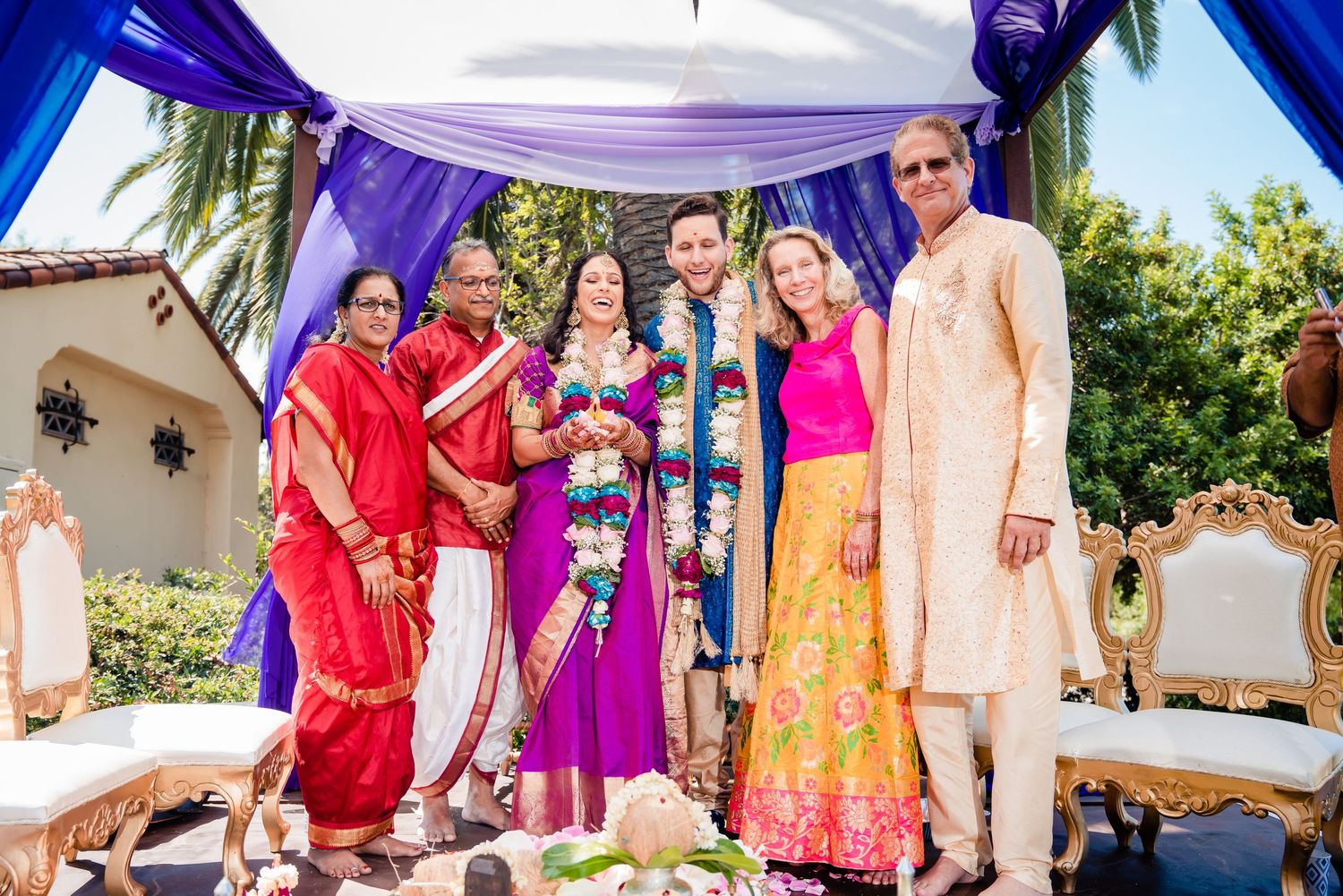 Colorfully dressed group posing together under a blue canopy at an outdoor celebration with palm trees in background.