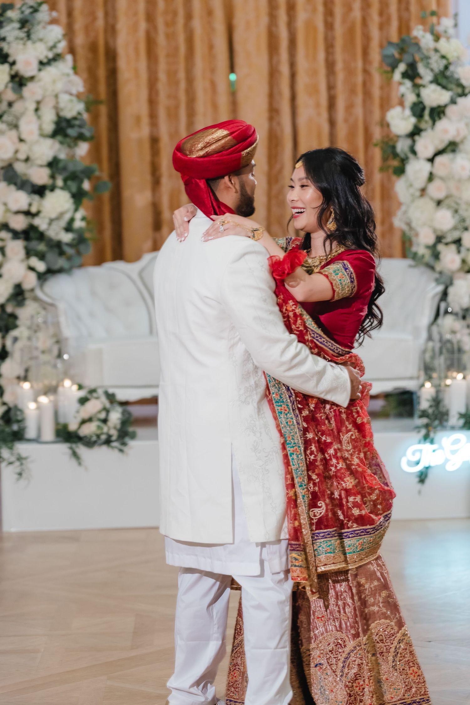 A couple in traditional South Asian wedding attire dance together in an elegant venue decorated with white floral arrangements.