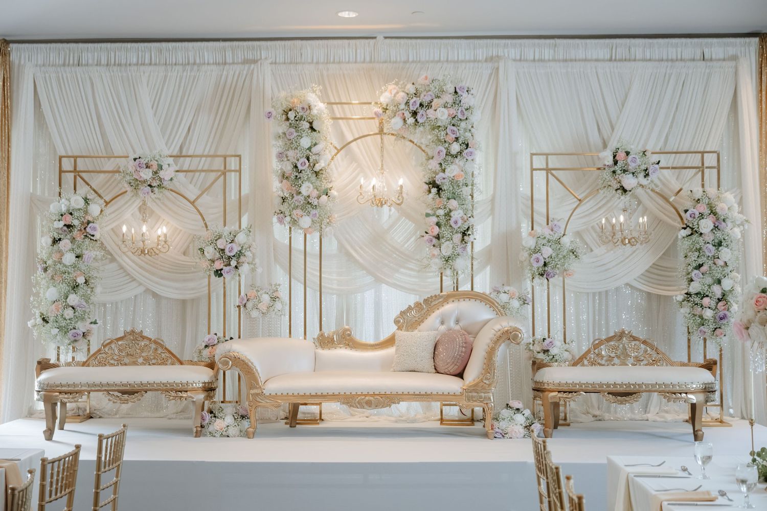 Elegant wedding stage with ivory chaise lounge, gold geometric backdrop, and white floral arrangements in a luxurious ballroom.