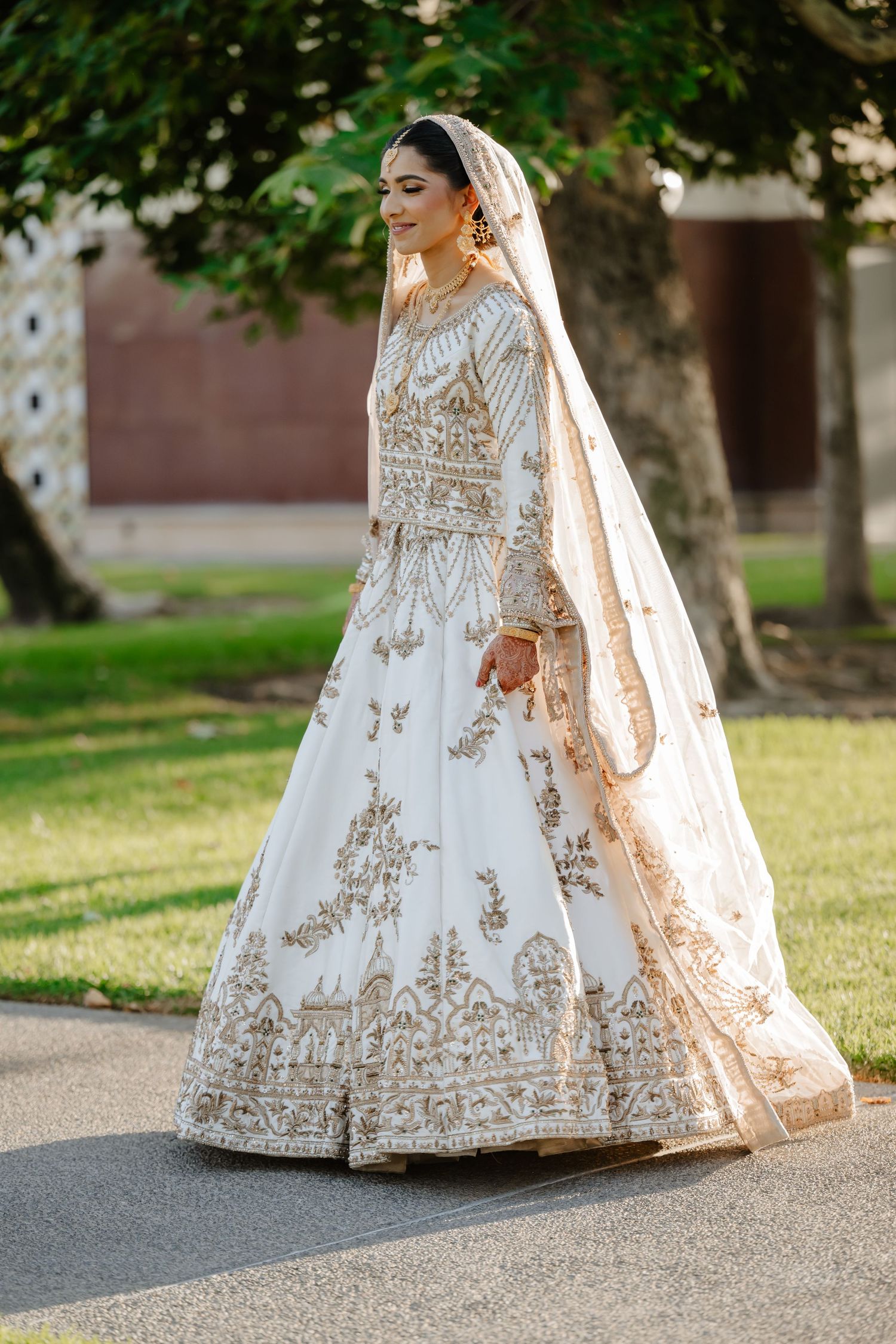 Bride in white and gold embroidered lehenga with dupatta walks outdoors on a sunny day.