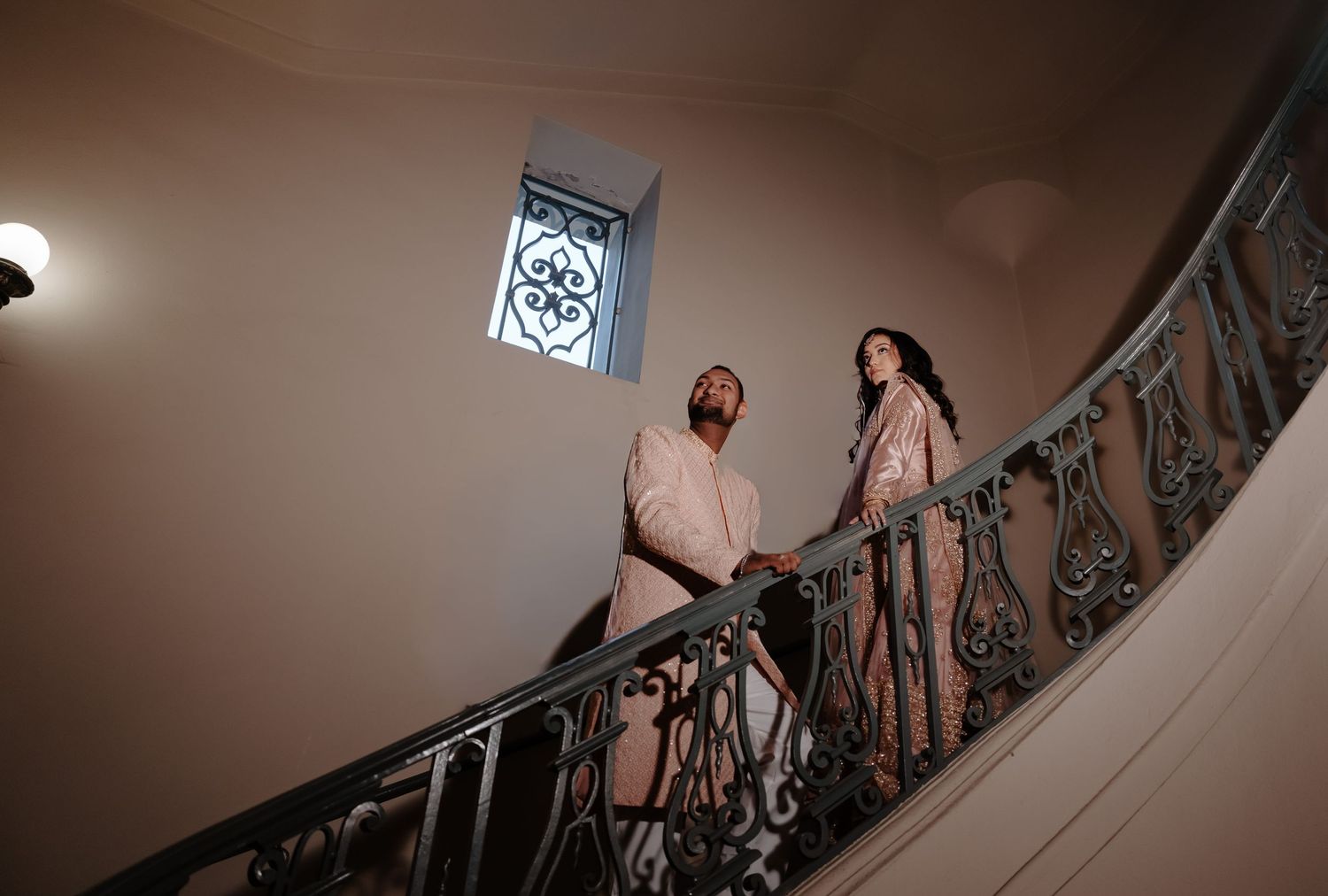 Two people in formal attire stand on an elegant curved staircase with ornate railings under dramatic lighting.