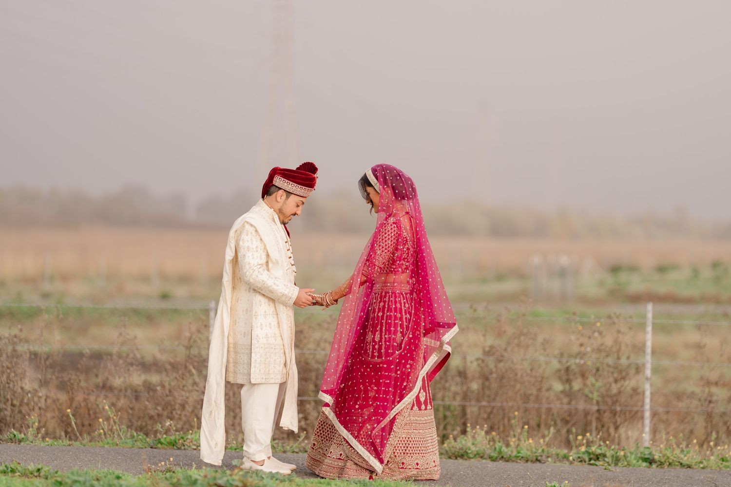Couple in traditional Indian wedding attire stands in a misty field during golden hour.