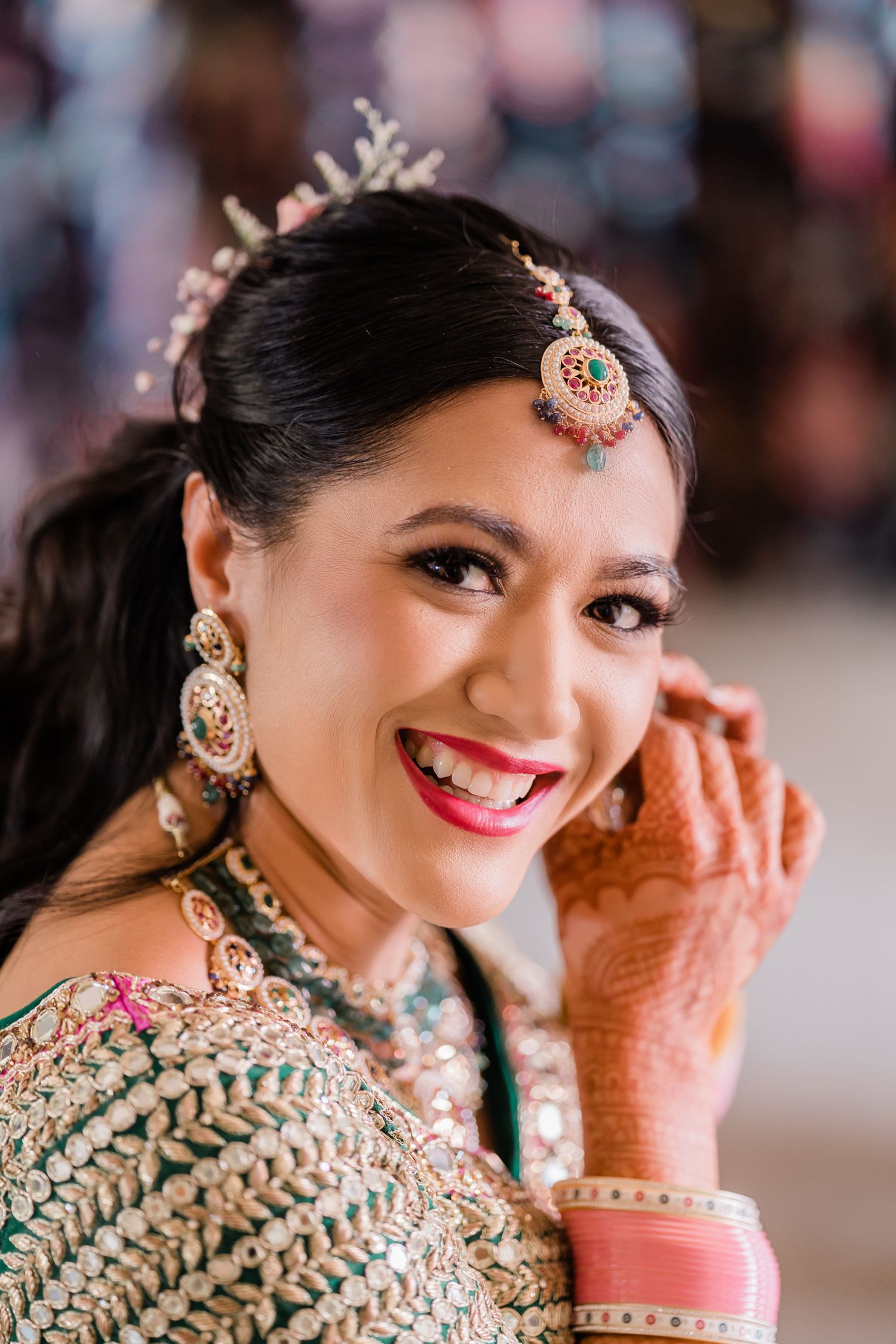 A bride in traditional Indian wedding attire smiles while wearing ornate jewelry and mehndi designs on her hands.