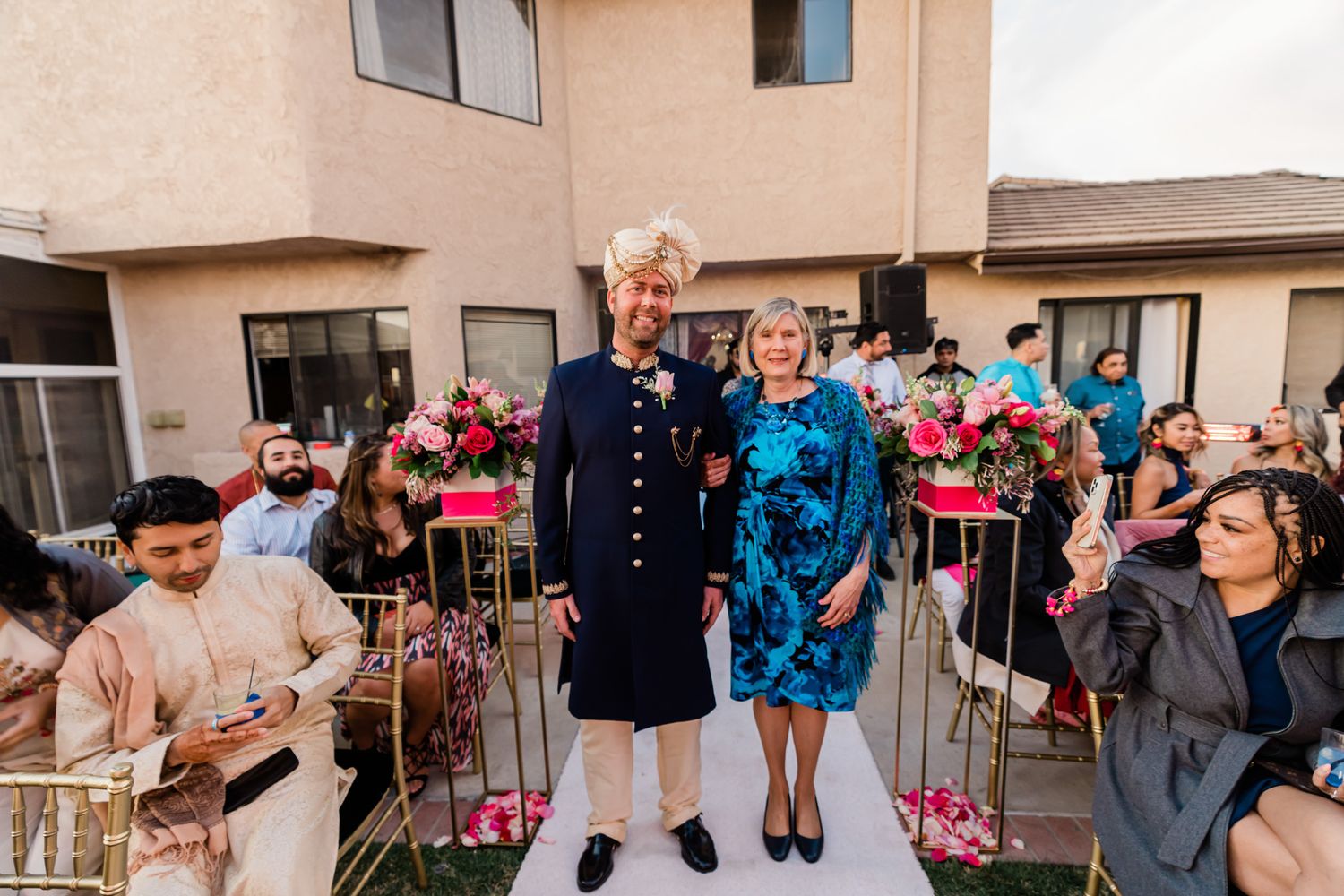 Outdoor celebration scene with guests gathered at a beige stucco building decorated with pink accents.