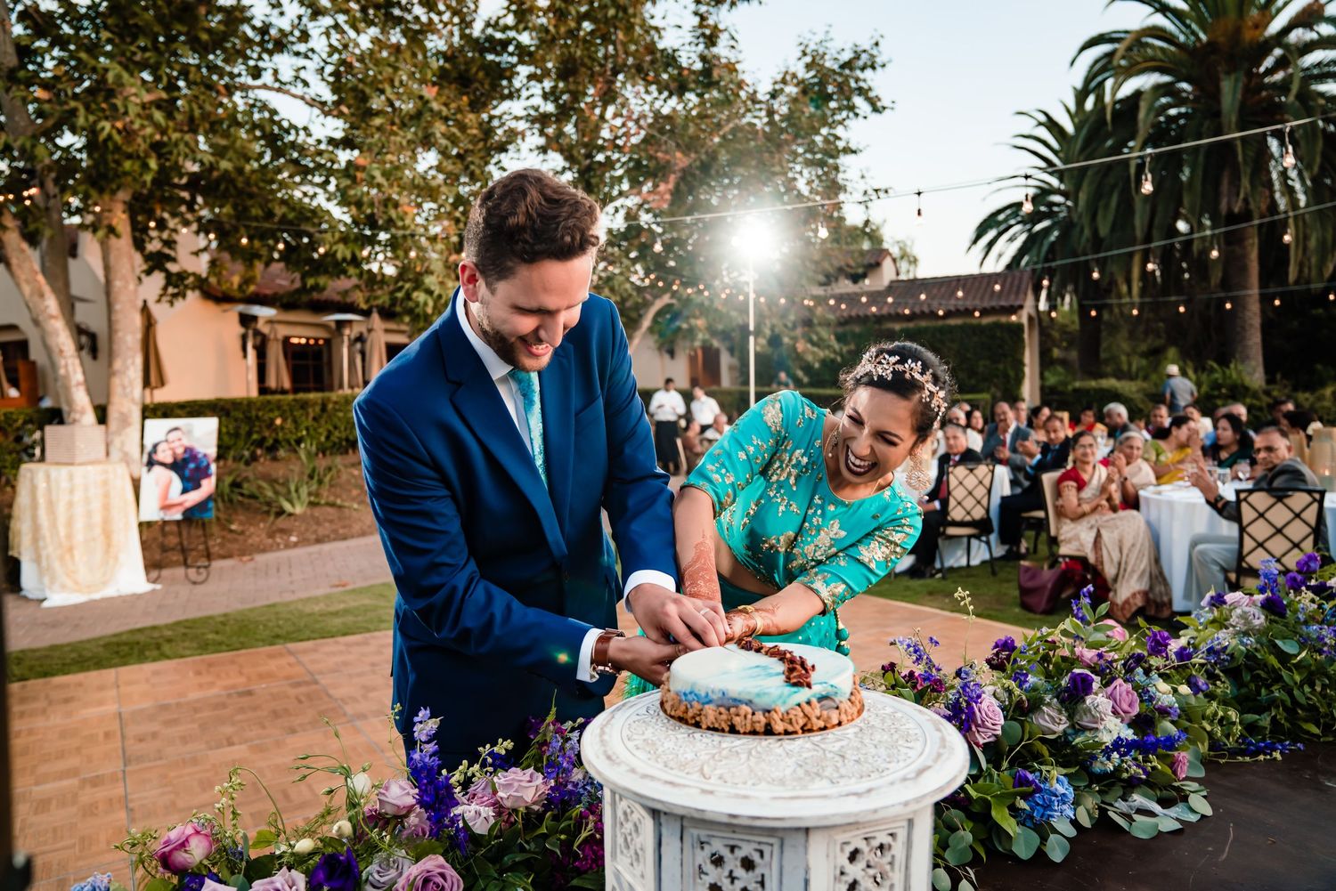 A wedding celebration outdoors with guests seated in rows and purple floral decorations along the aisle.