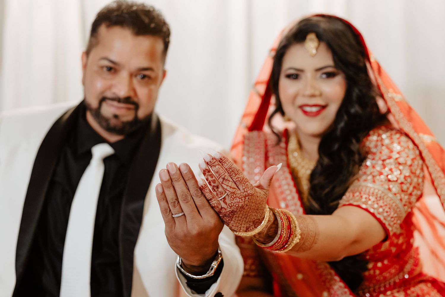 Indian bride in red lehenga showing mehndi hands with groom in black suit at traditional wedding ceremony.