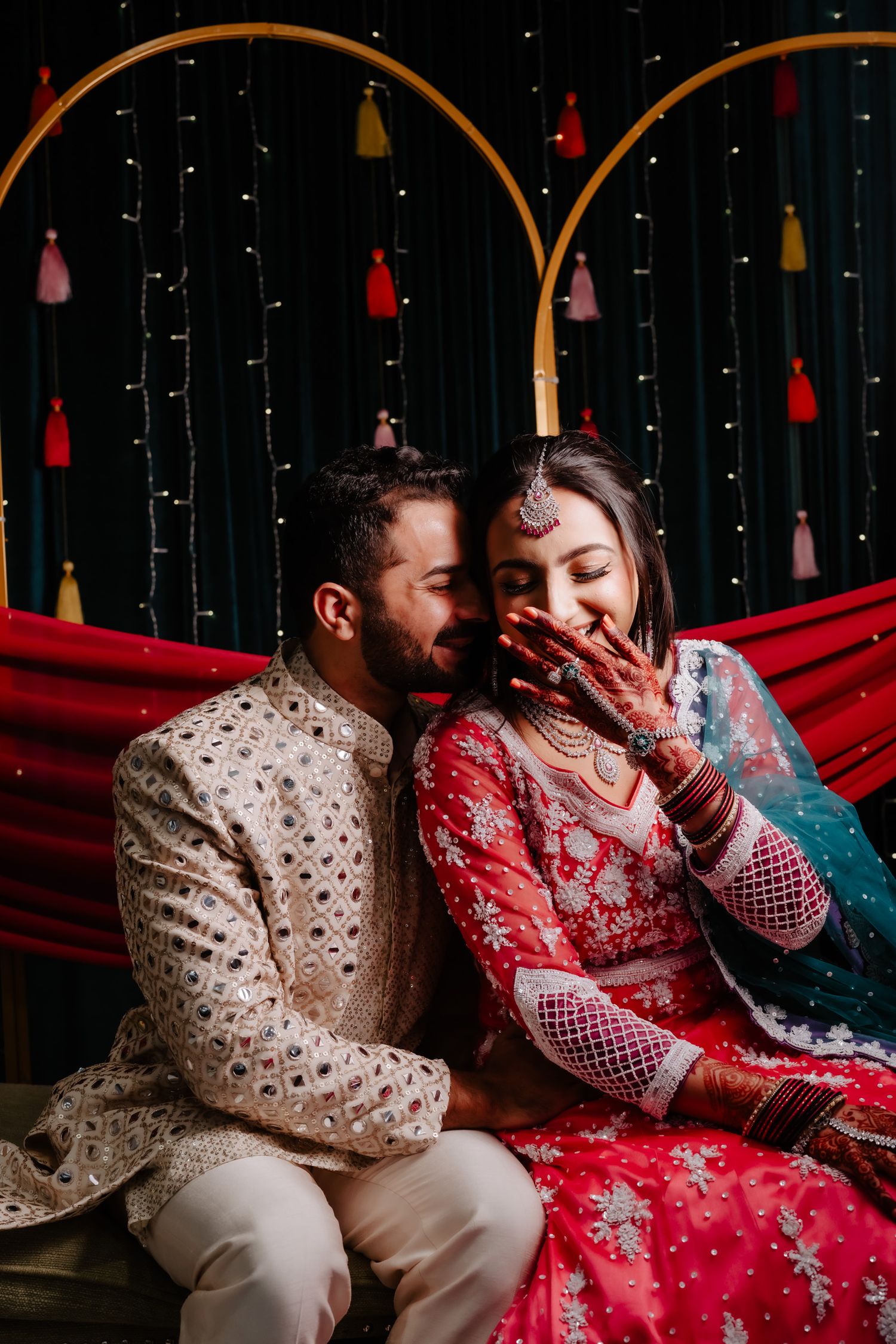 A couple in traditional South Asian attire share romantic moments in a dimly lit setting with hanging lights and tassels.