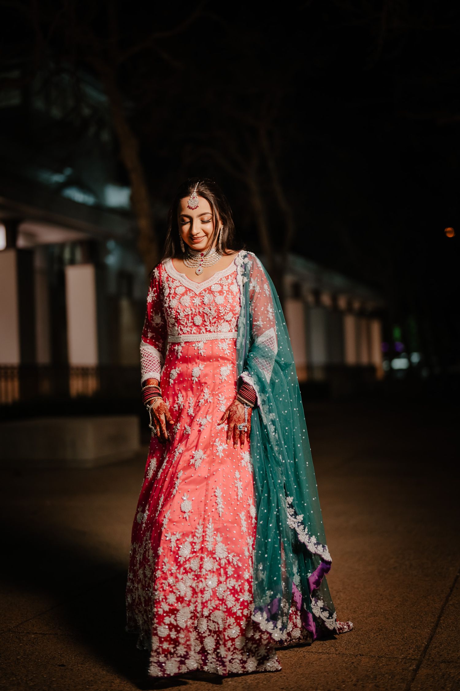 A person wearing a coral pink floral embroidered lehenga with a teal dupatta poses in a dimly lit outdoor setting.