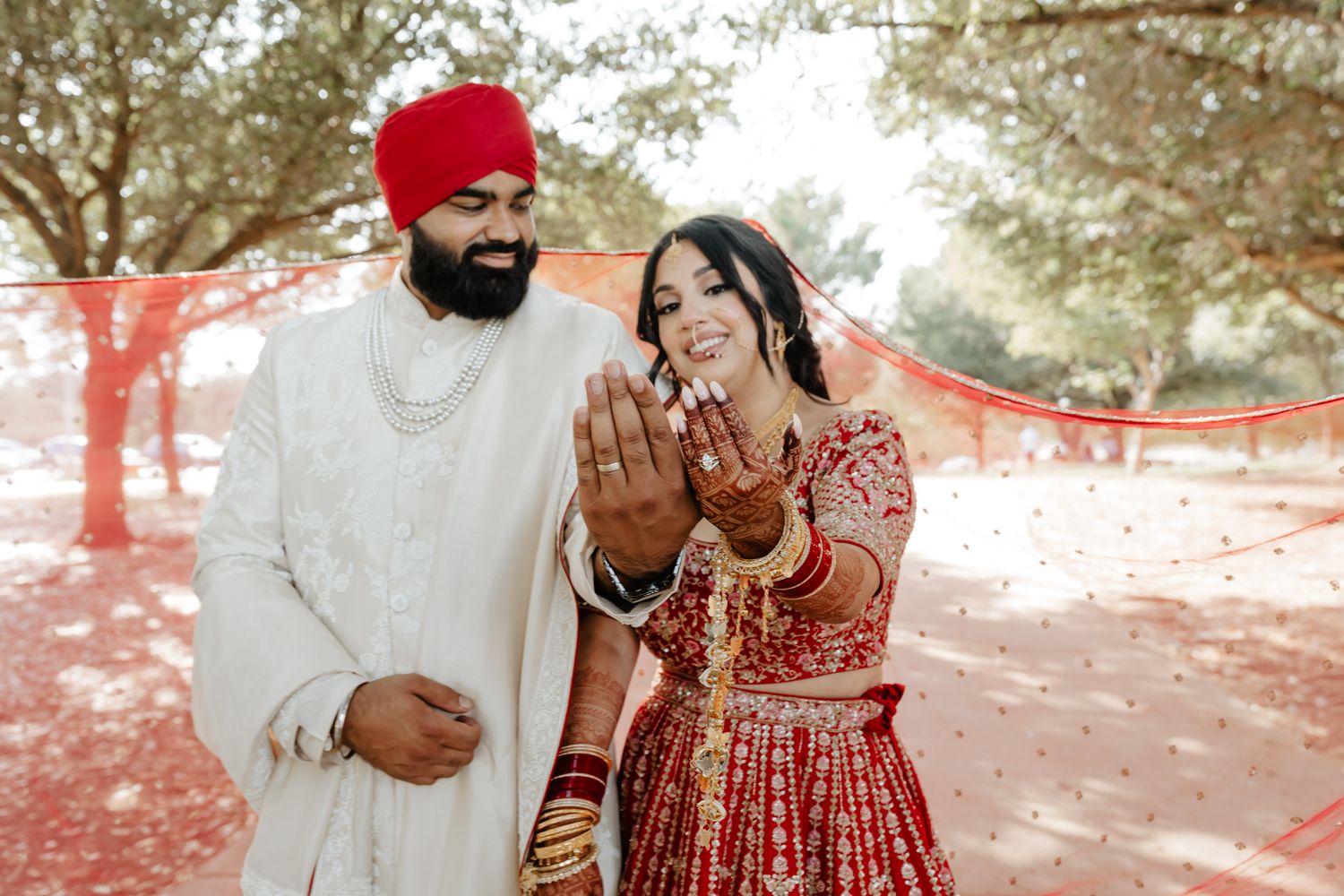 A Sikh couple in traditional wedding attire share a joyful moment outdoors with the bride showing her henna-adorned hands.