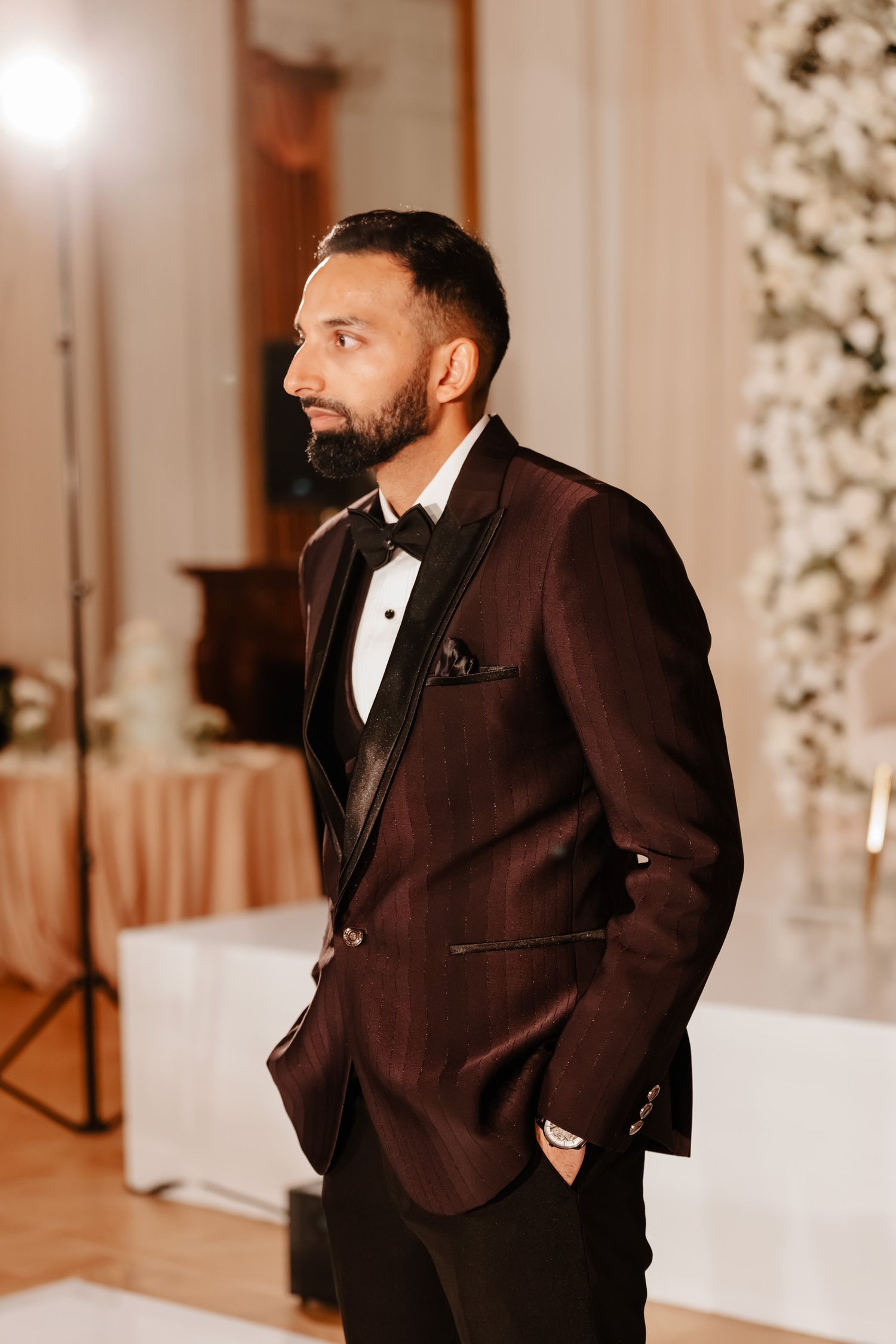 Groom in a classic black tuxedo poses during wedding reception with elegant white floral backdrop.