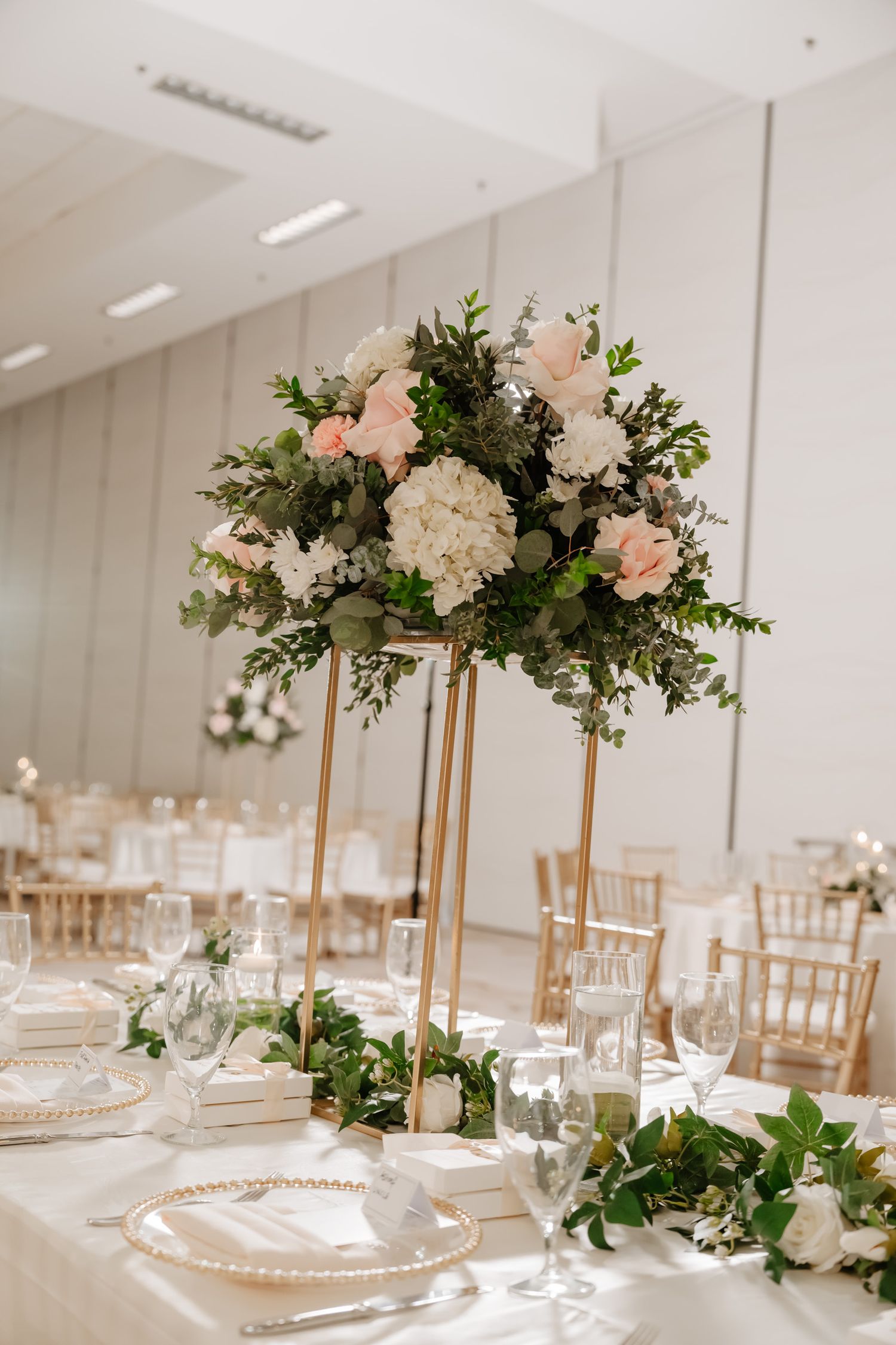 Modern white wedding reception venue with tall floral centerpieces and gold chiavari chairs.