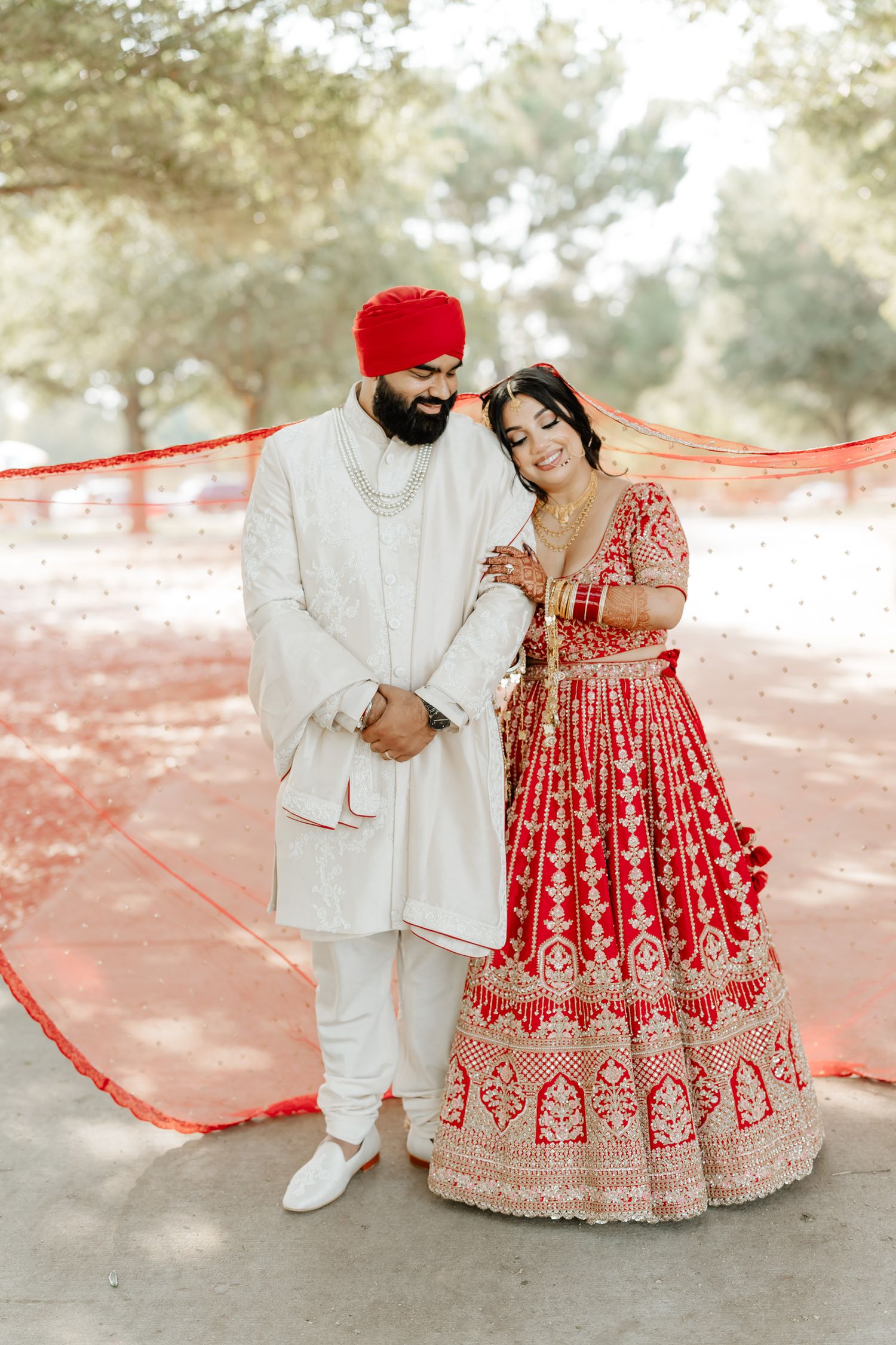 A couple in traditional Indian wedding attire pose together against a dreamy outdoor backdrop with red decorations.