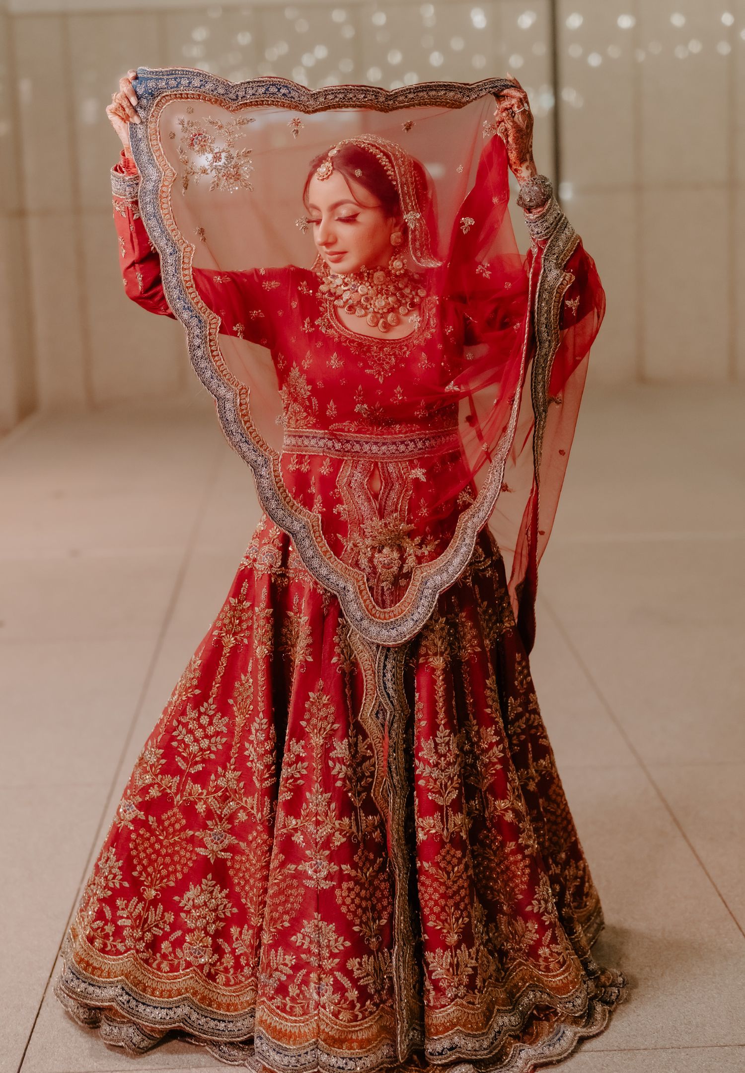 Traditional Indian bridal lehenga in vibrant red with intricate gold embroidery and sheer veil displayed against neutral backdrop.