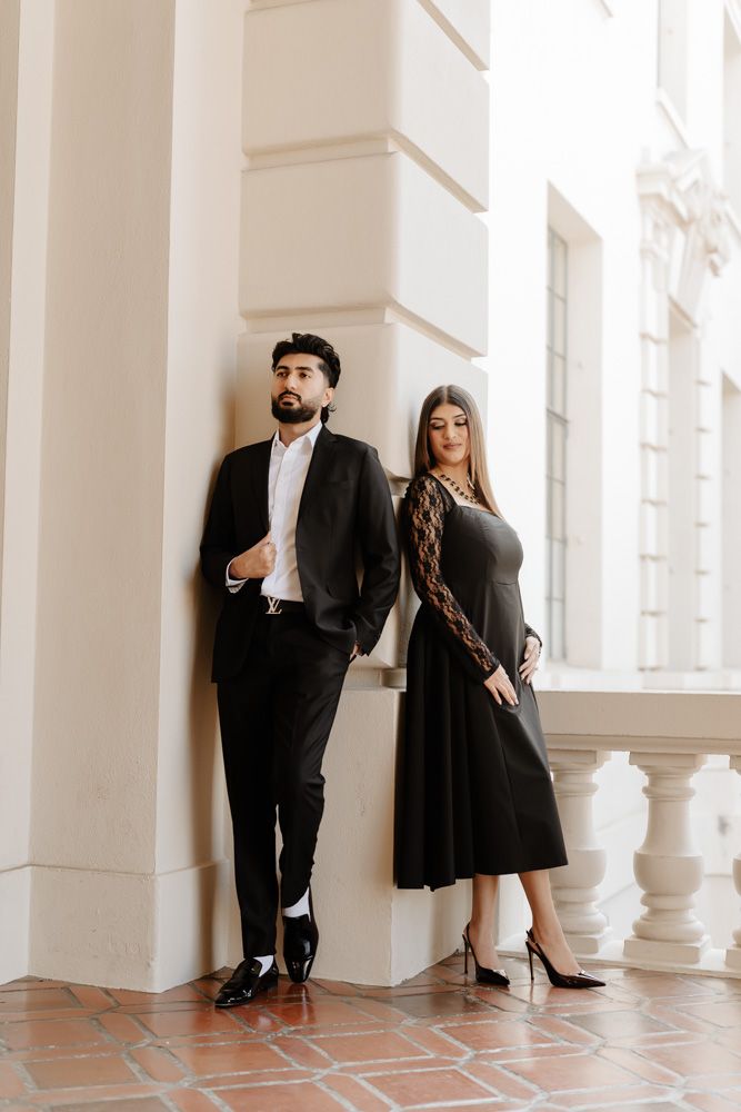 Elegant couple in matching black formal attire posing against white architectural columns.