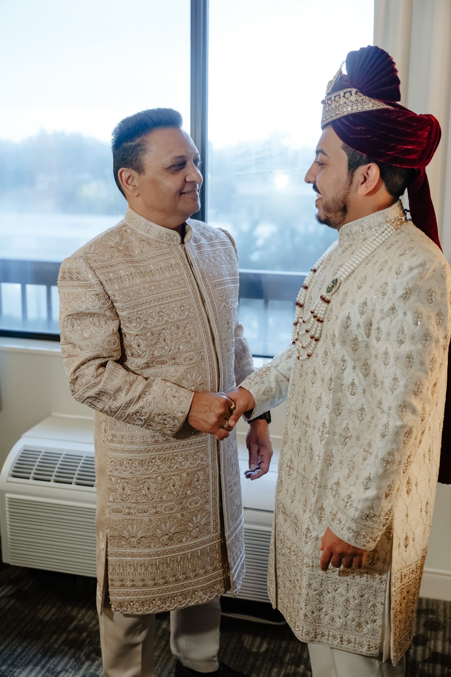 Two people in traditional embroidered formal attire interact near a window in an indoor setting.