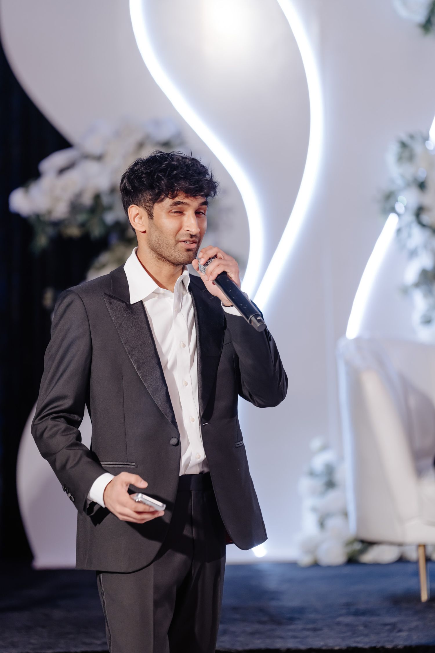 A man in a dark suit checks his phone against a white backdrop with LED lighting.