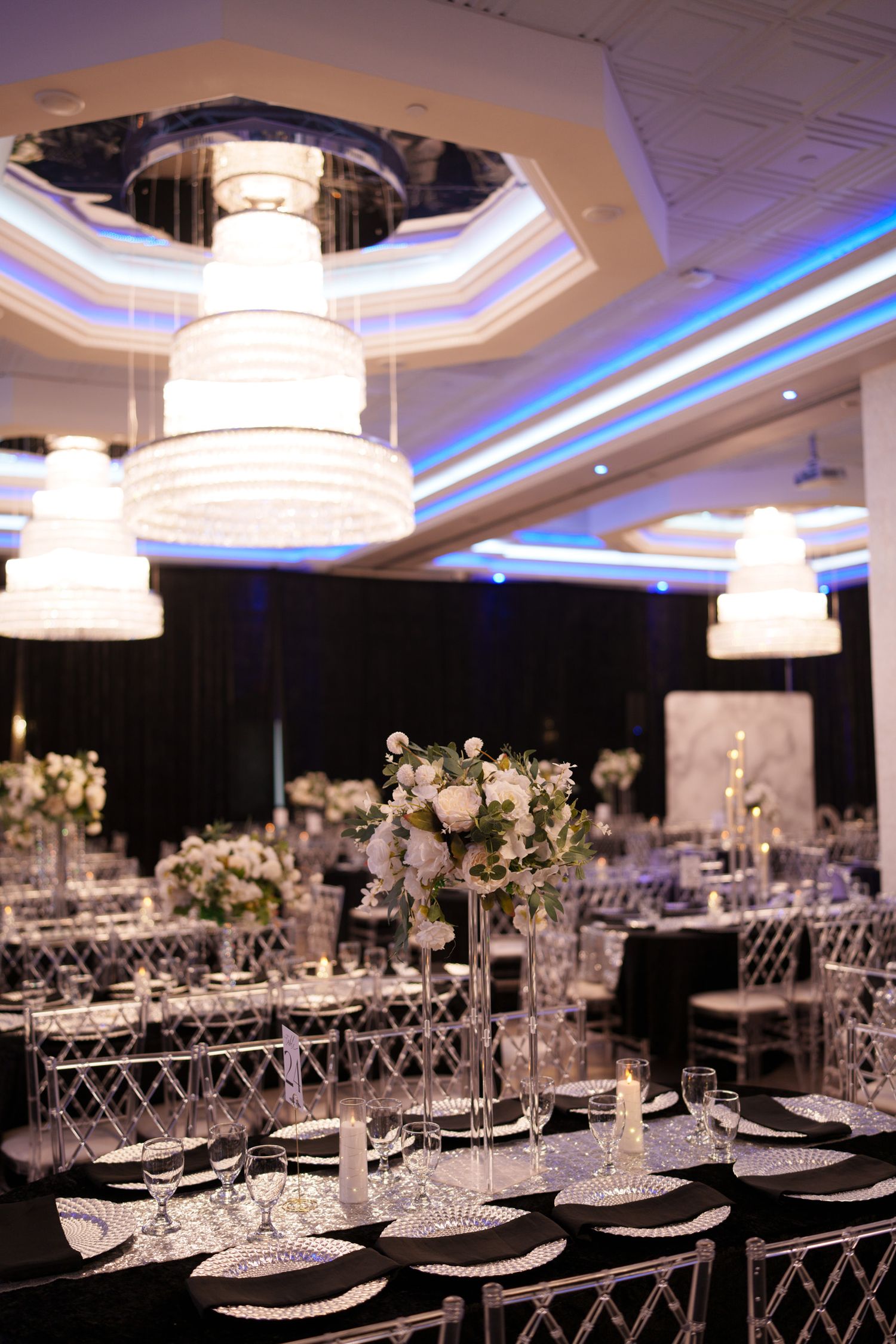Elegant wedding reception venue with crystal chandeliers, white floral centerpieces, and modern table settings in blue lighting.