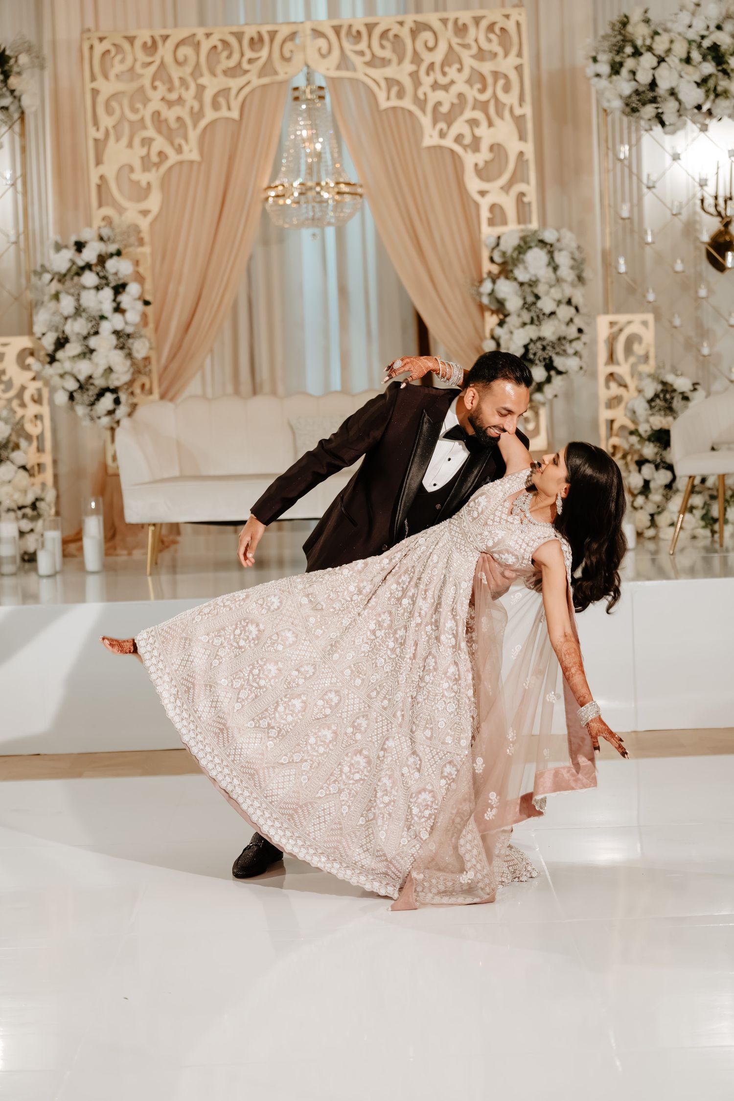 Couple performs romantic dance dip pose on white dance floor with ornate backdrop and floral decorations.
