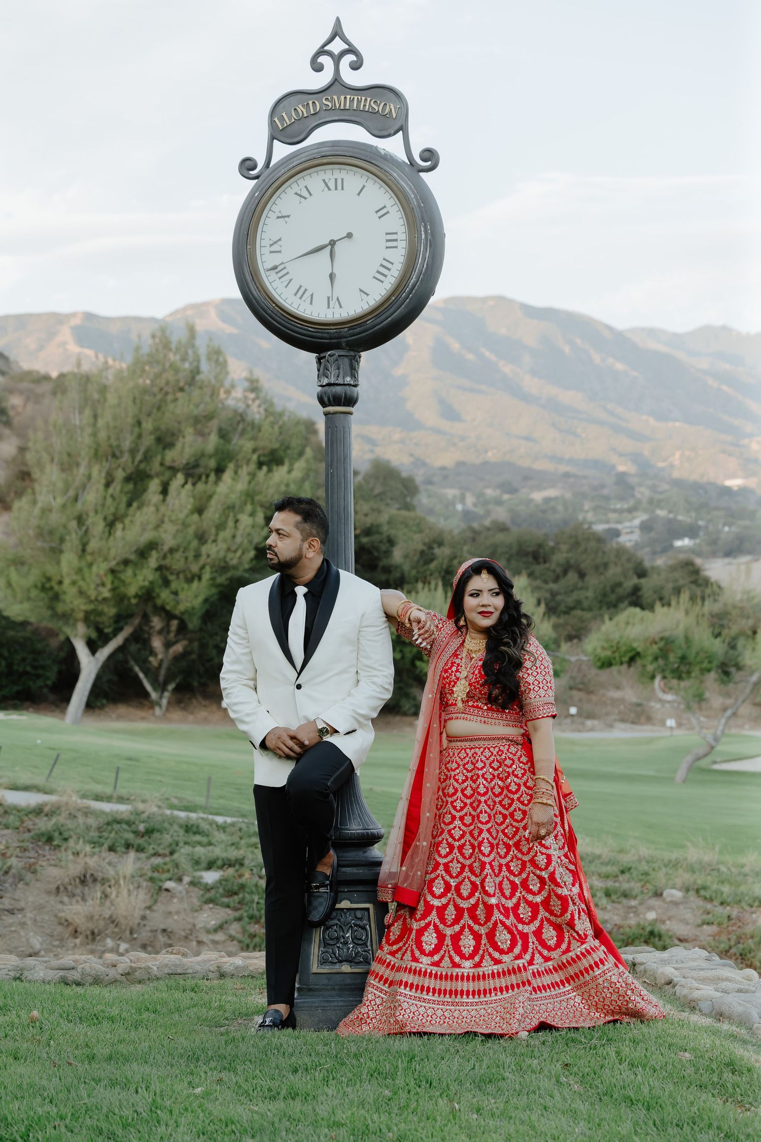 Indian bride in red lehenga and groom in white tuxedo pose by a clock tower with mountain backdrop.