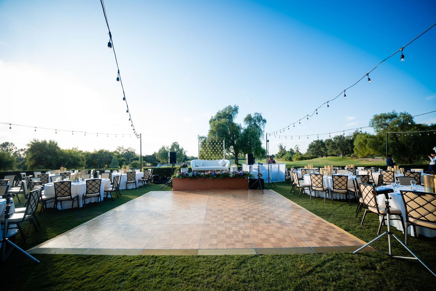 An outdoor wedding reception setup with a wooden dance floor, string lights, and white chairs arranged on grass under blue sky.