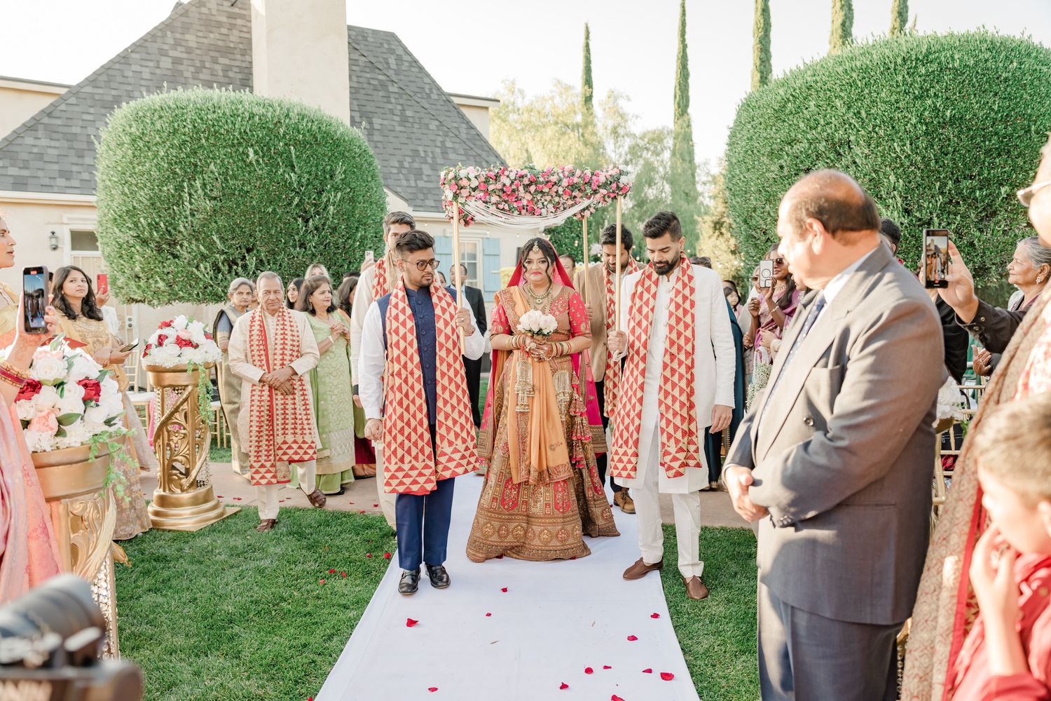 Outdoor wedding ceremony with guests in colorful attire surrounded by manicured hedges and cypress trees.