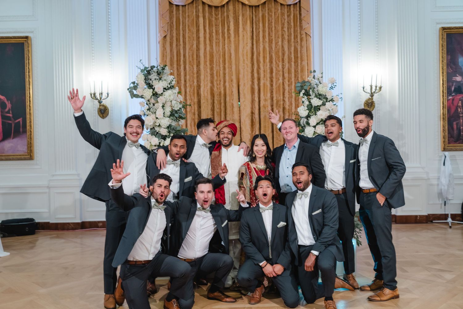 A group of people in formal attire posing playfully in a grand white room with elegant decor.