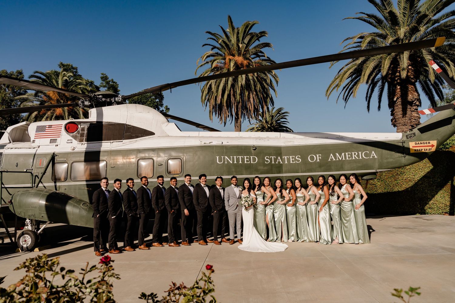 Wedding party poses in front of palm trees and ocean backdrop at an elegant outdoor venue.