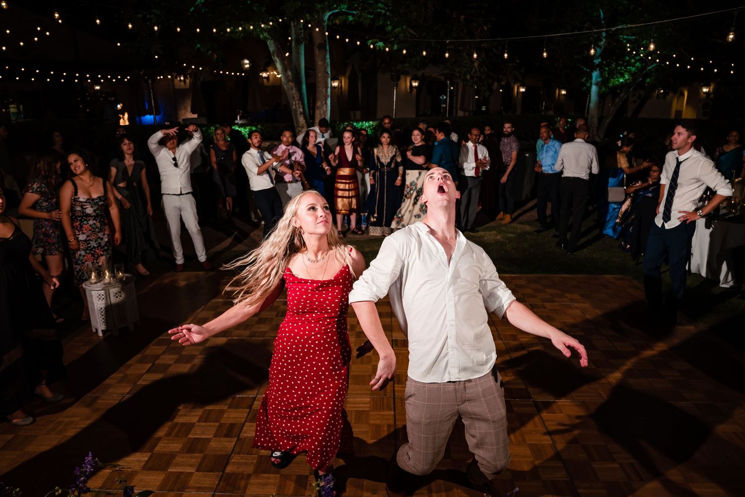 A group of people dancing and celebrating at a dimly lit nighttime outdoor event with string lights overhead.