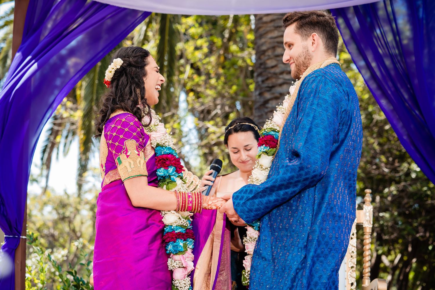A couple in bright pink and blue outfits share a romantic moment during an outdoor photoshoot in a palm tree setting.