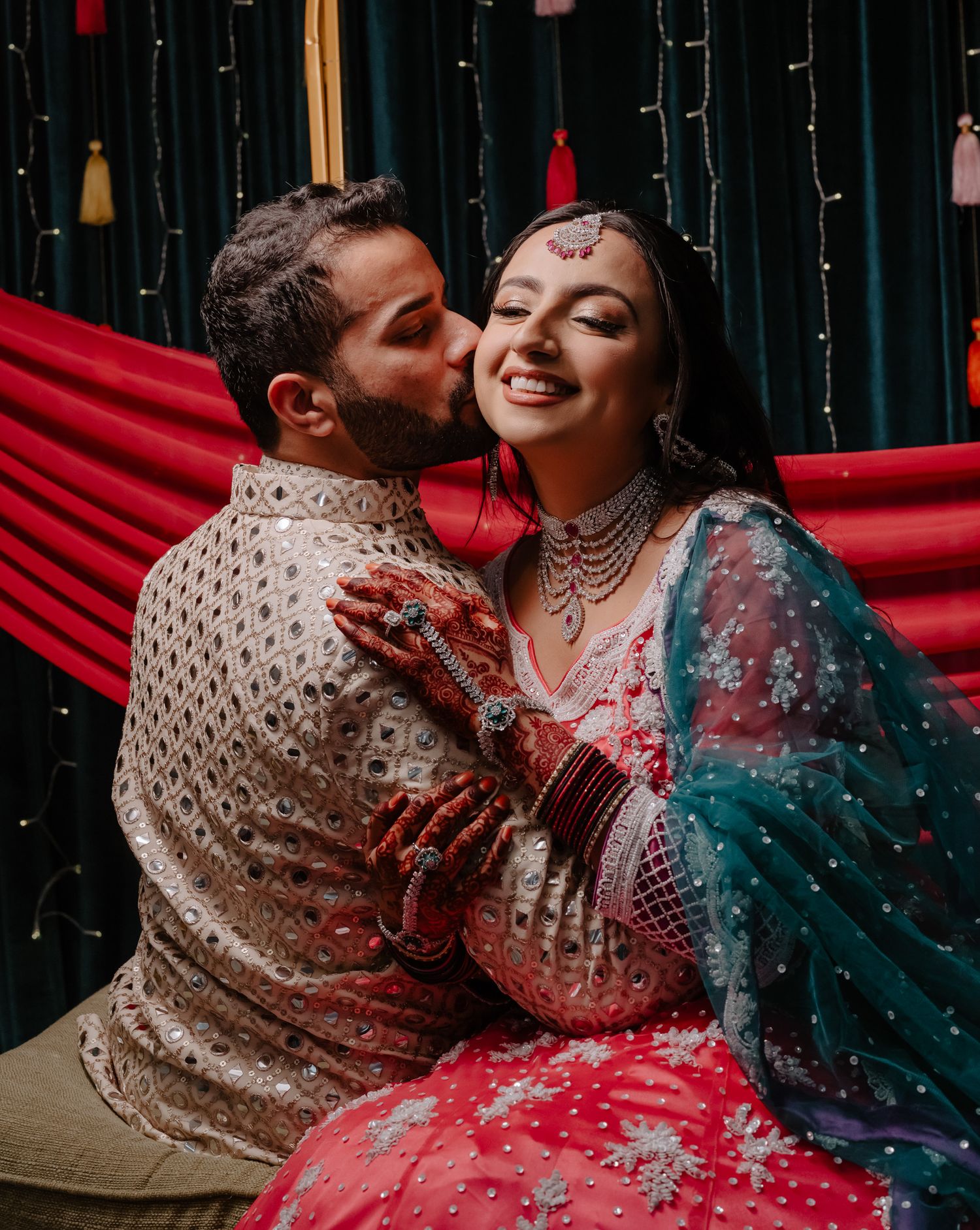 A couple in ornate South Asian wedding attire shares tender moments against a backdrop with red drapery.