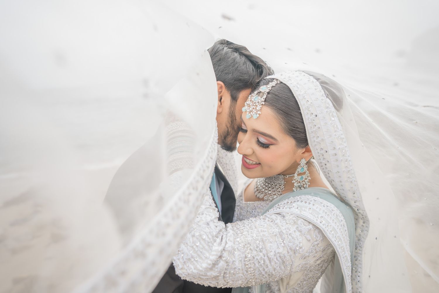A bride in an elegant white lace wedding dress and flowing veil poses gracefully in a romantic setting.