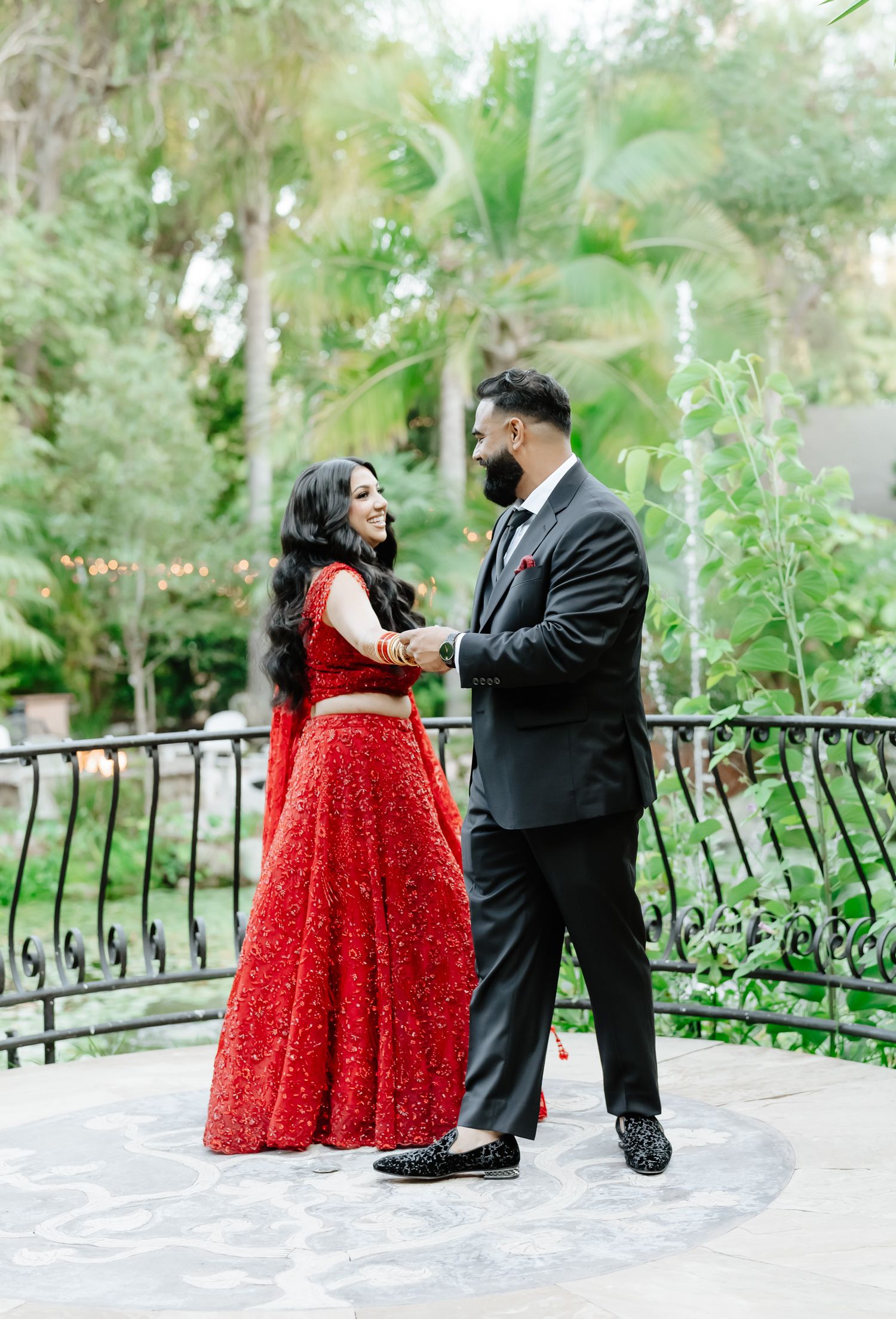 A couple in formal attire shares a romantic moment in a lush garden setting with palm trees.