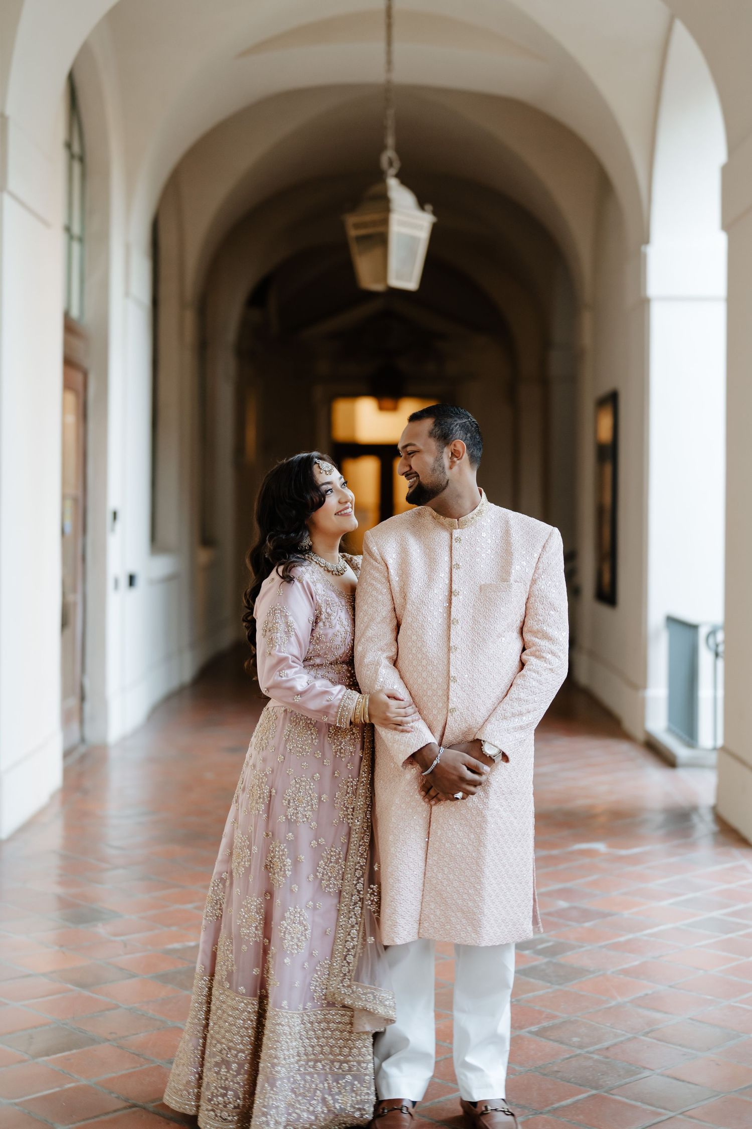 A couple in traditional South Asian wedding attire stands lovingly in an arched hallway.