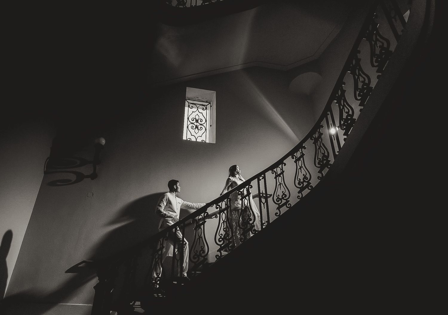 A person stands on an elegant curved staircase with ornate iron railings in dramatic black and white lighting.
