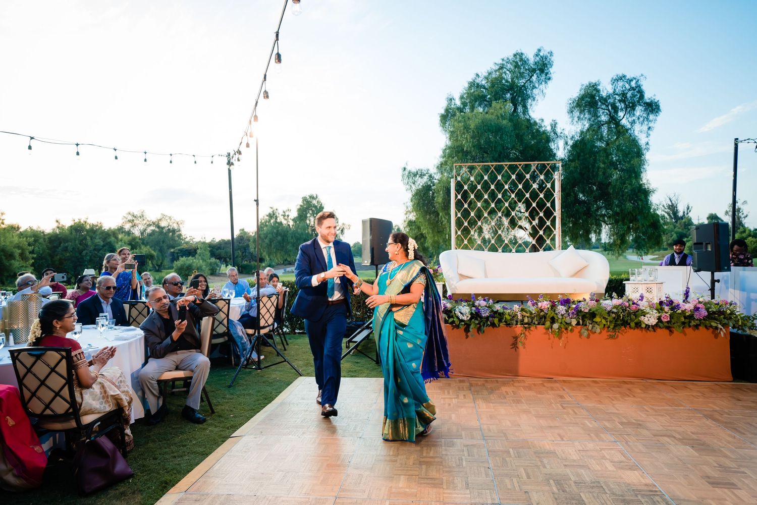 Outdoor wedding reception setup with string lights and lattice decorations during golden hour.