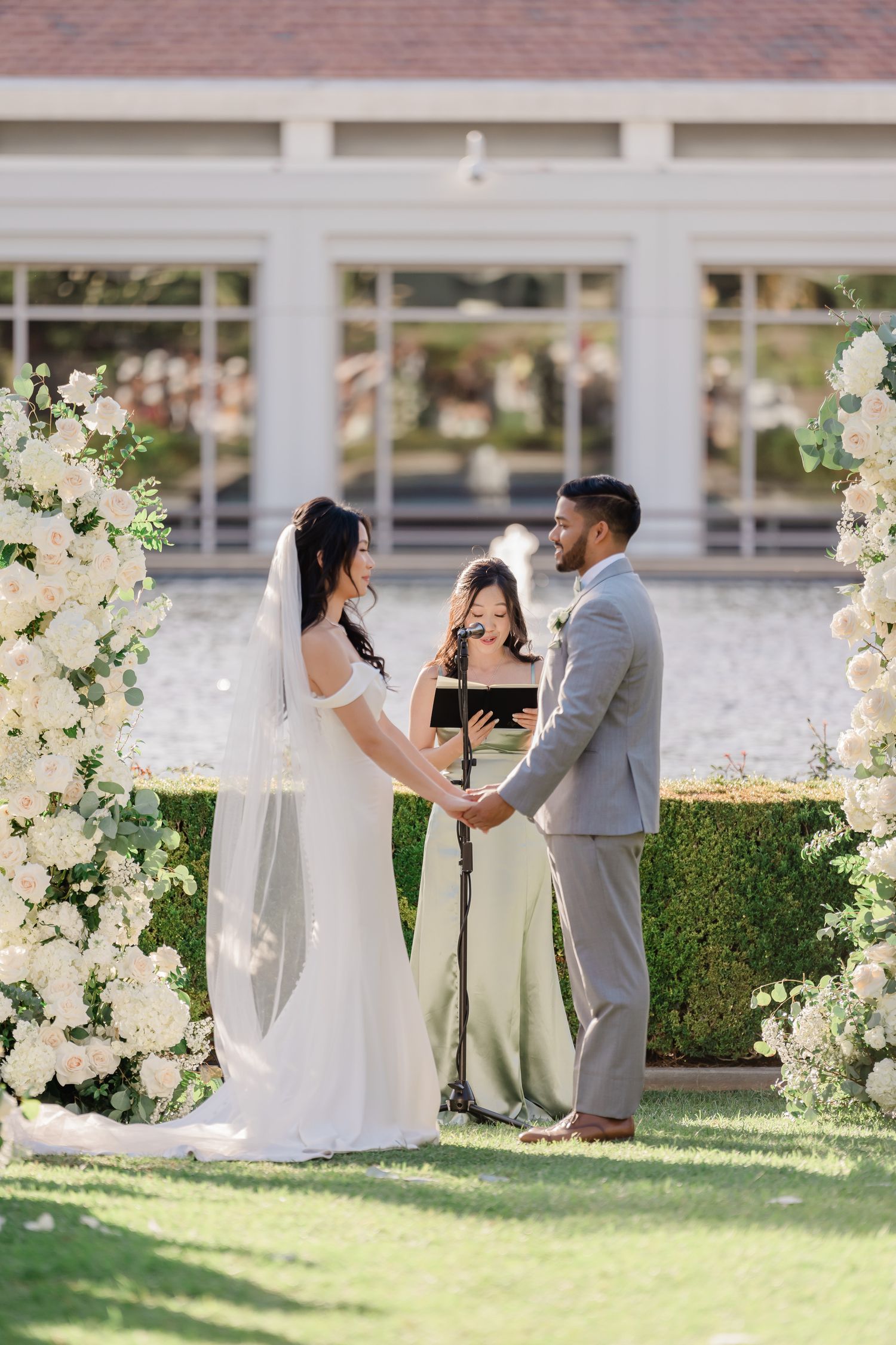 Wedding couple poses for photos among white floral arrangements and elegant garden columns.